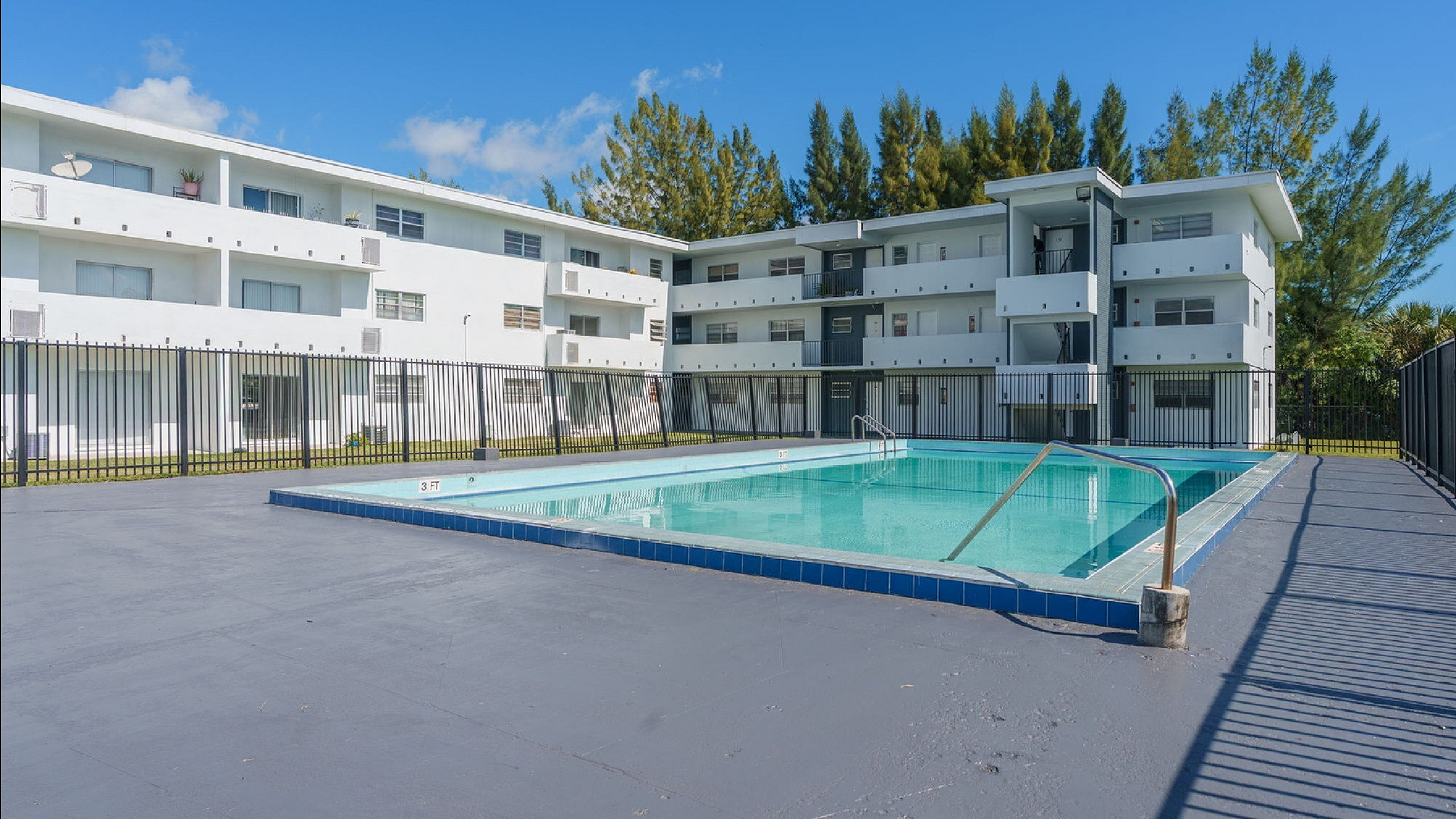 Sunlit apartment complex with a sparkling outdoor pool, surrounded by modern architecture and lush greenery.