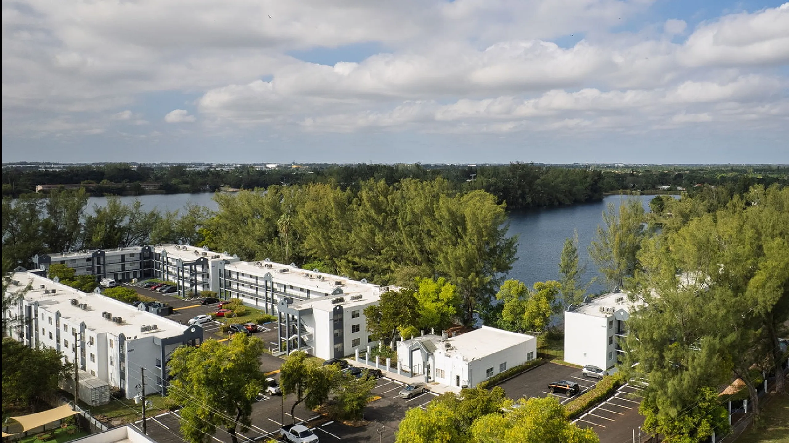 Stunning aerial view of modern apartment complex surrounded by lush greenery and overlooking a serene lake under a bright blue sky.