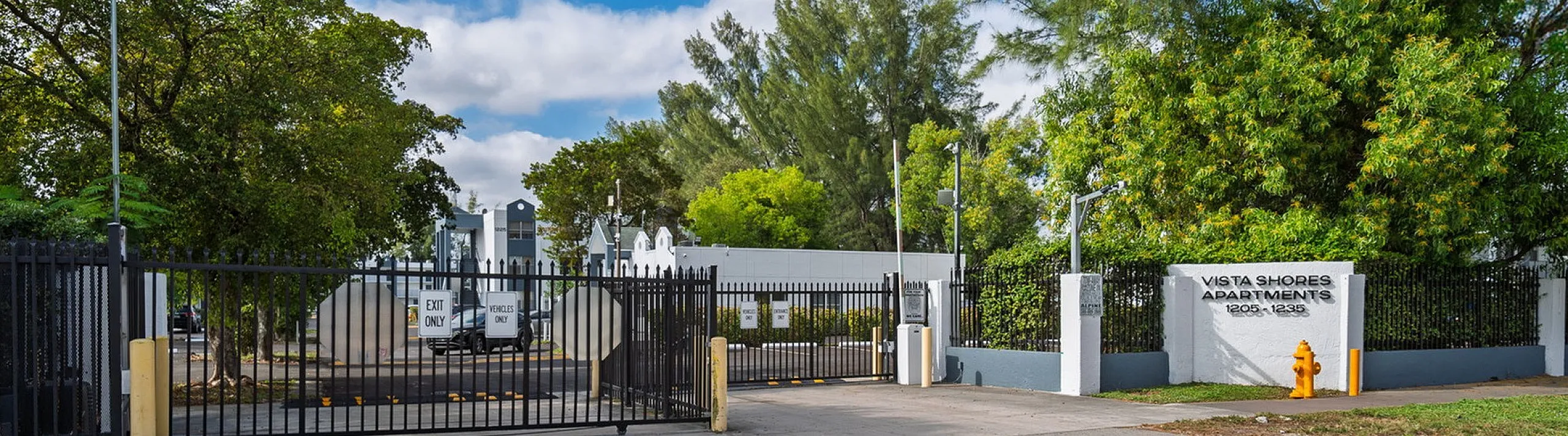 Welcoming entrance of Vista Shores Apartments surrounded by lush greenery and modern security gates under a clear blue sky.