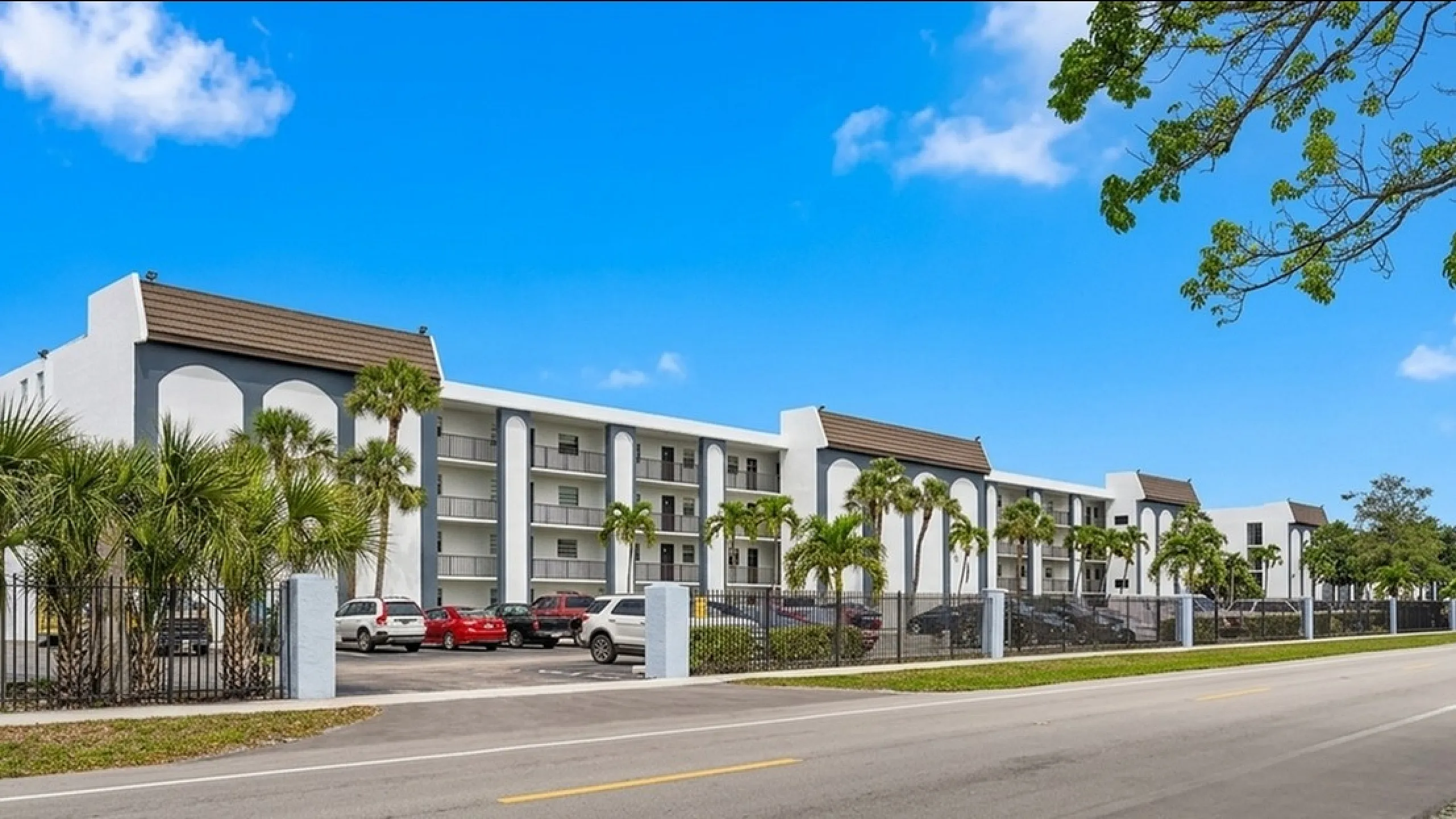 Contemporary apartment complex with palm trees and ample parking, set against a bright blue sky, promising relaxed living and convenience.