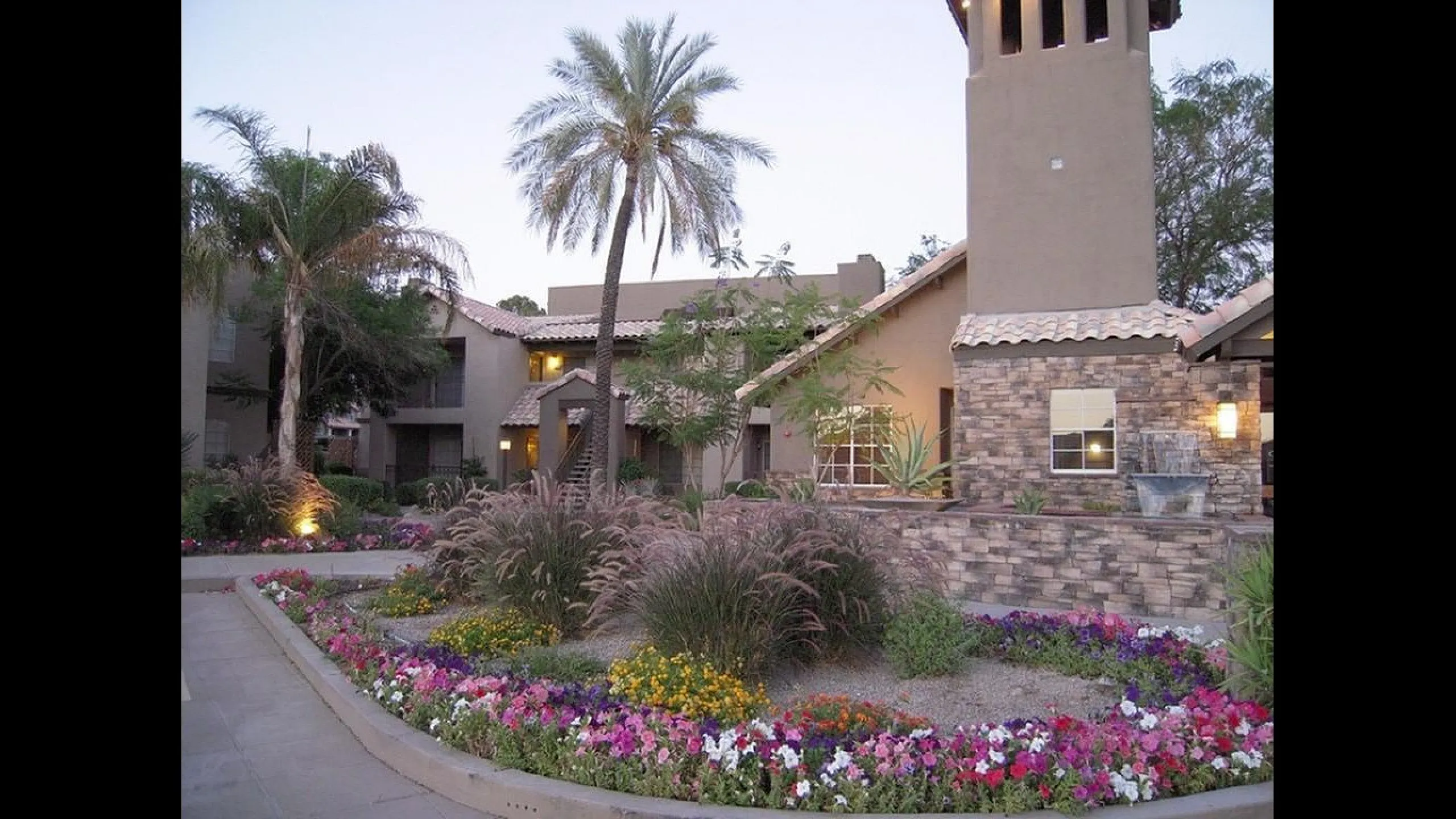 Charming apartment exterior with rustic stone facade, lush palm trees, and vibrant flower gardens creating a serene and inviting ambiance.