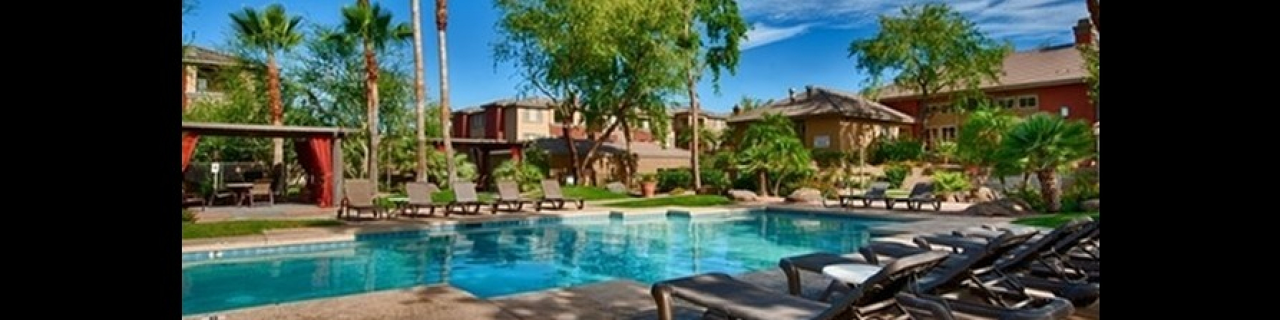 Luxurious apartment community pool area with lush landscaping, comfortable lounge chairs, and vibrant palm trees under a clear blue sky.