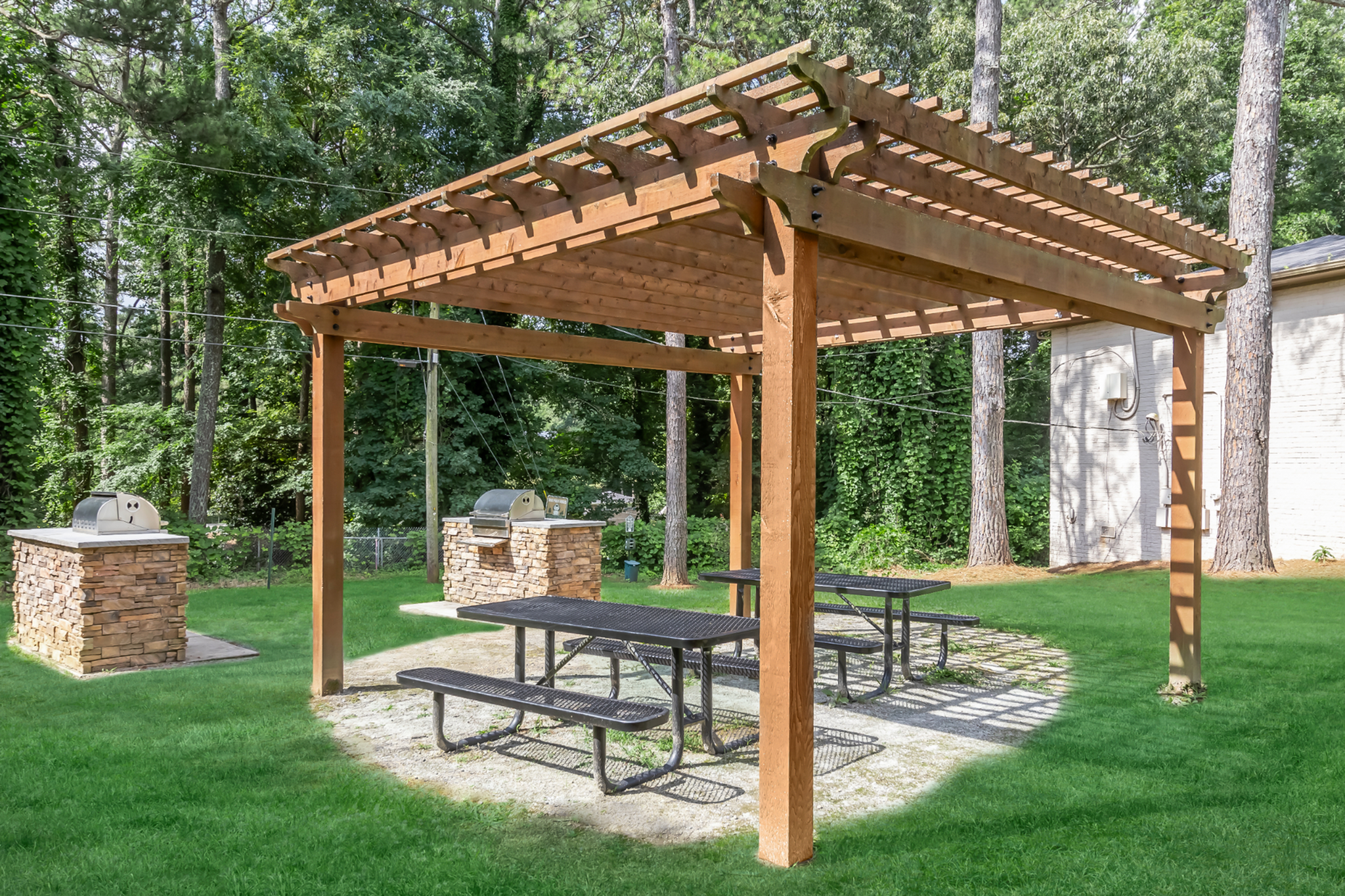 Charming outdoor space featuring a wooden pergola with picnic tables and built-in stone grills, surrounded by lush greenery.