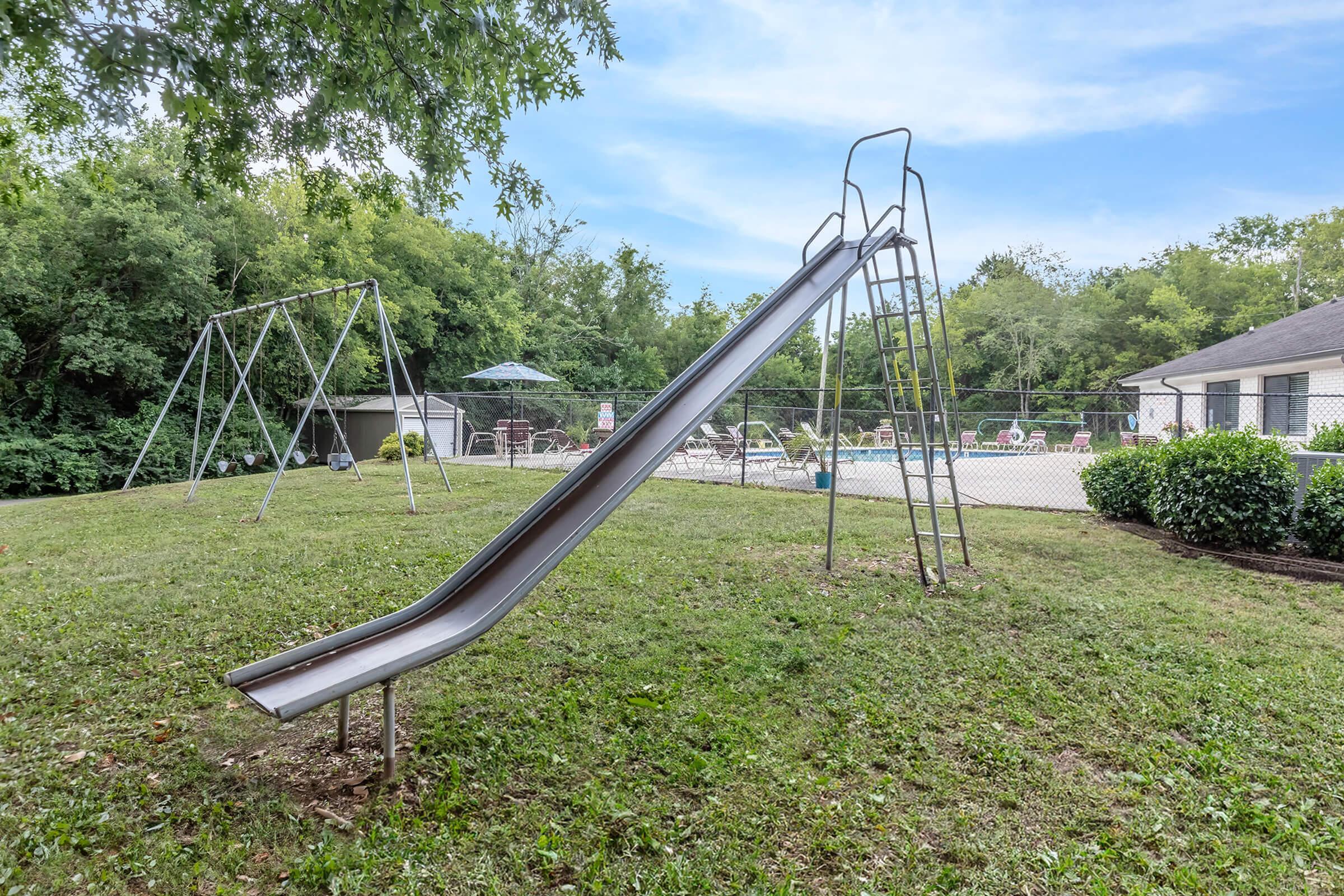 Charming outdoor playground with a slide and swings, surrounded by lush greenery, adjacent to a refreshing swimming pool area.