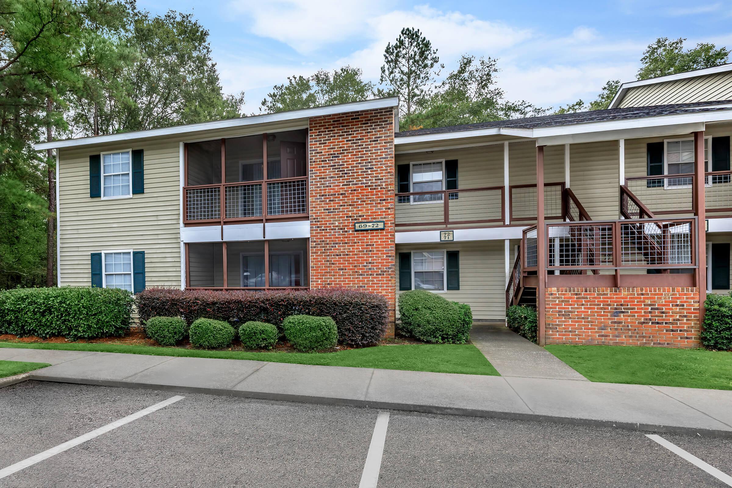 Charming two-story apartment building with brick and siding exterior, featuring lush greenery and ample parking in a serene neighborhood setting.