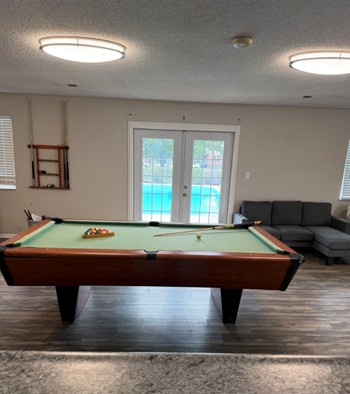 Modern recreation room featuring a stylish pool table, cozy seating area, and bright double doors opening to a refreshing pool view.