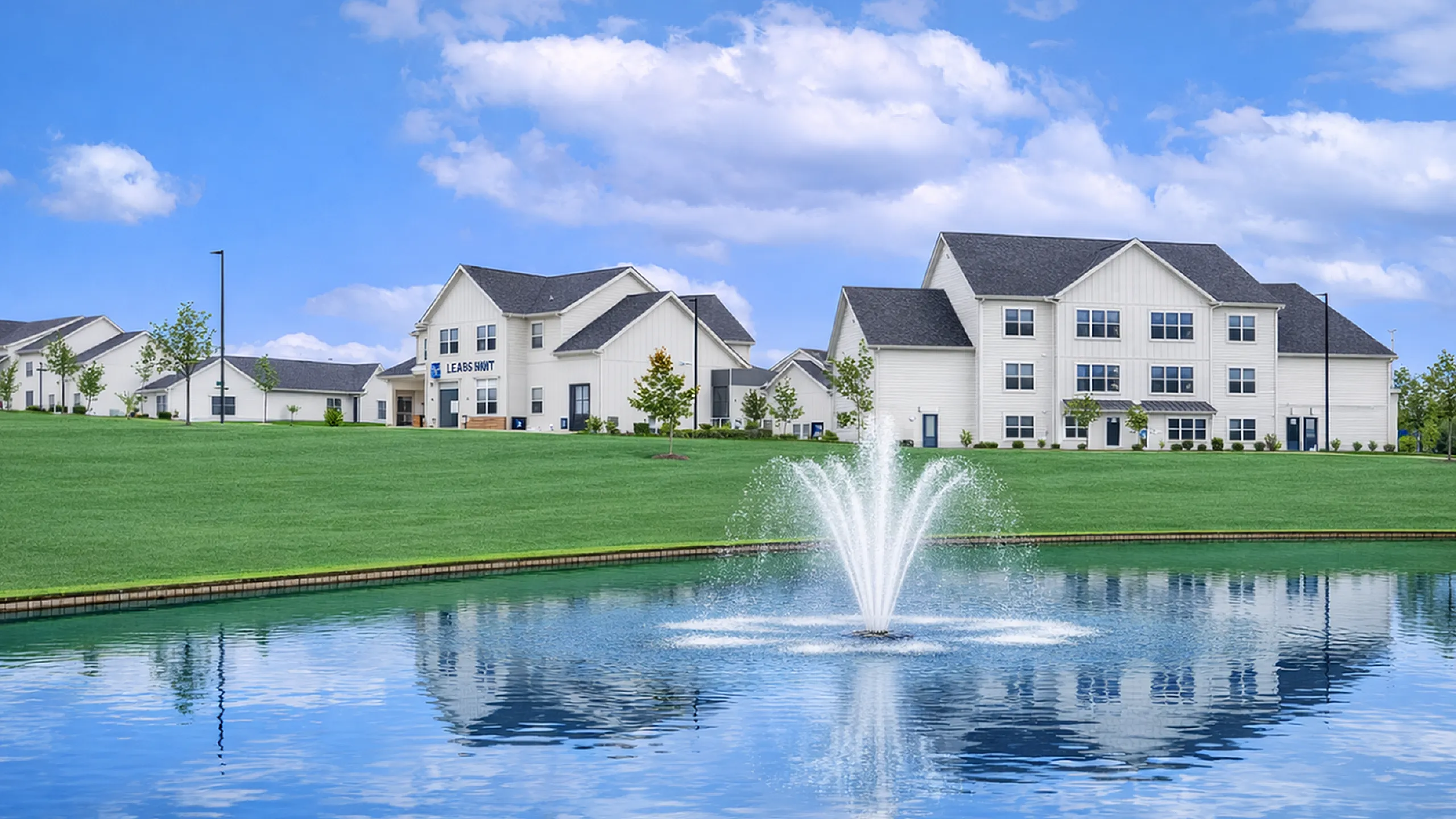 Charming apartment community with scenic pond and fountain view, nestled among lush green lawns under a bright blue sky.