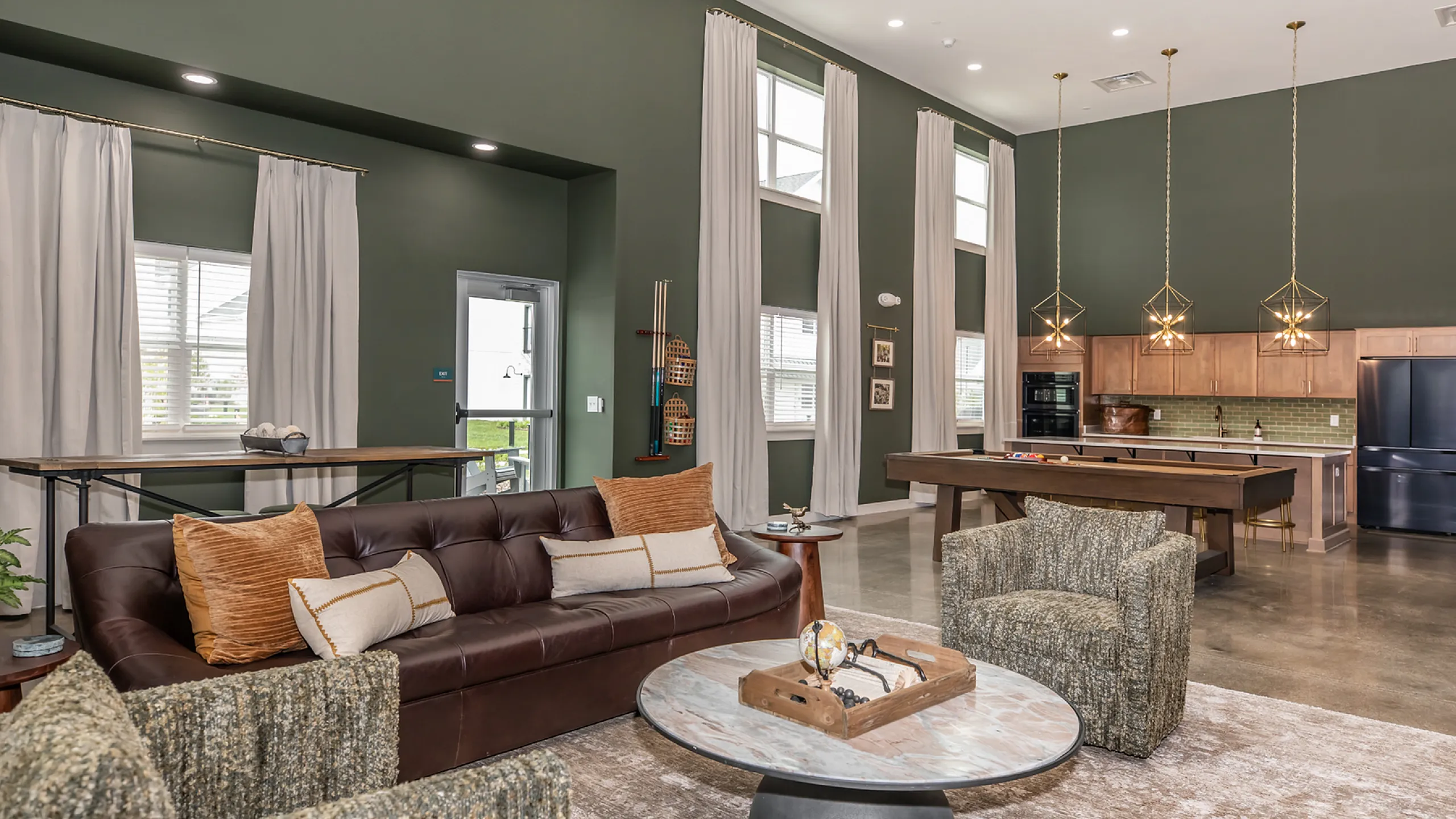 Chic living room with dark green walls, featuring a plush leather sofa, stylish armchairs, and a modern kitchen with elegant pendant lighting.