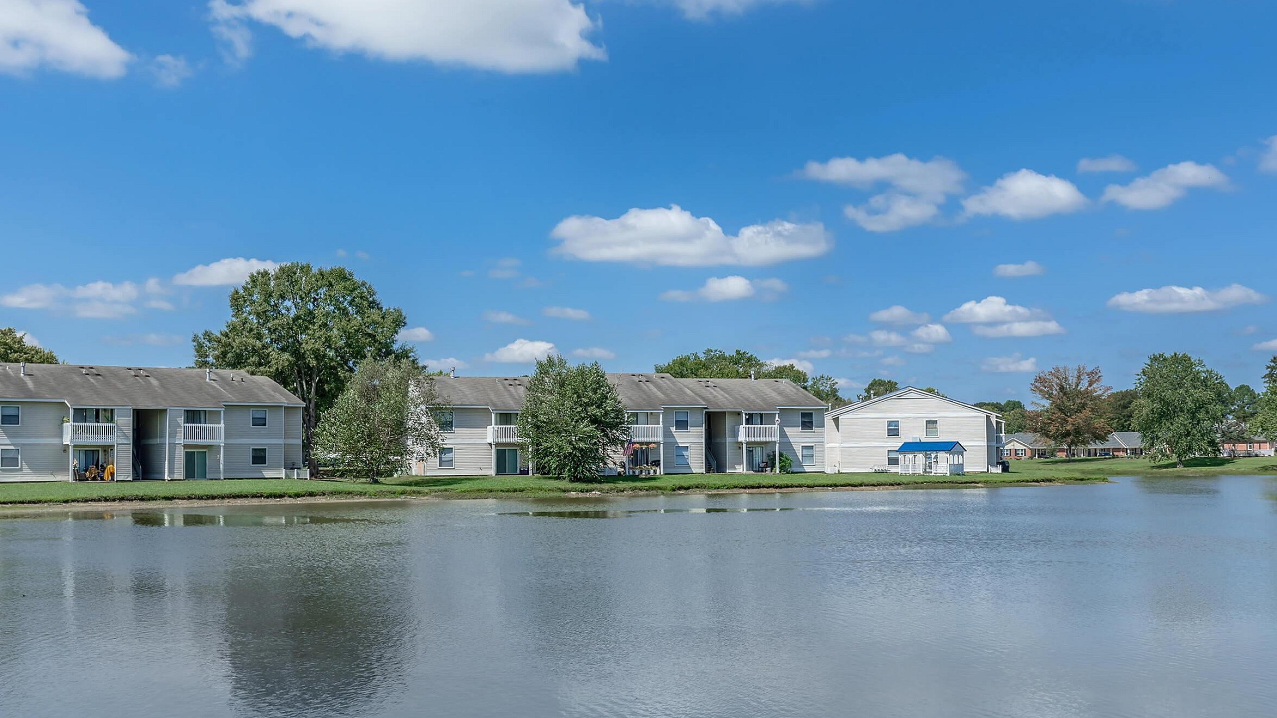 Serene lakeside view of charming white apartments with lush greenery and clear blue skies.
