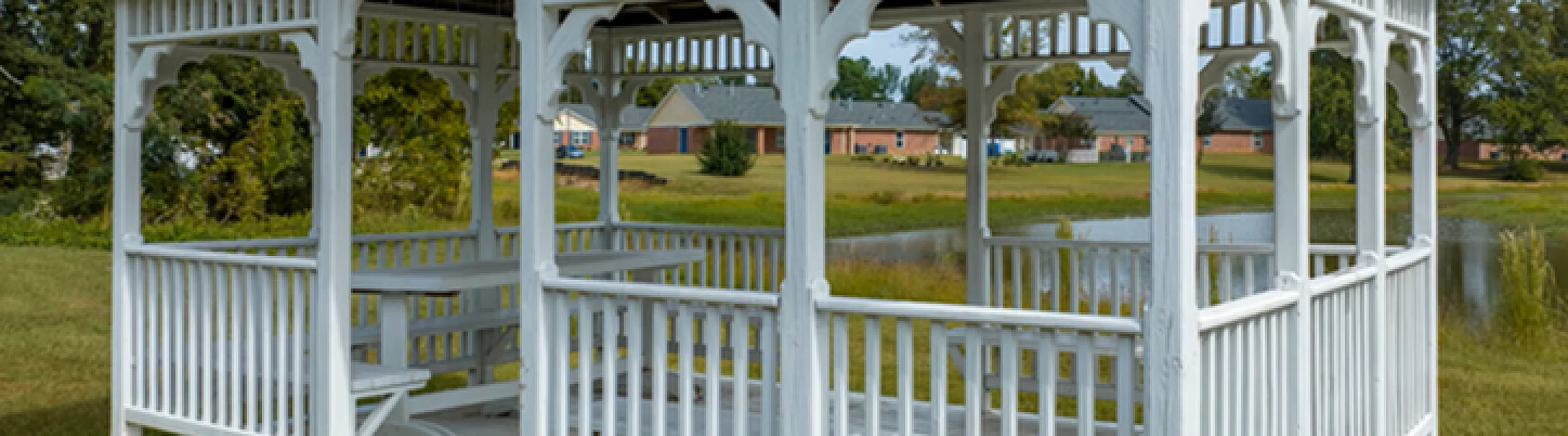 Charming white gazebo with a blue metal roof, surrounded by lush greenery and overlooking a tranquil pond.