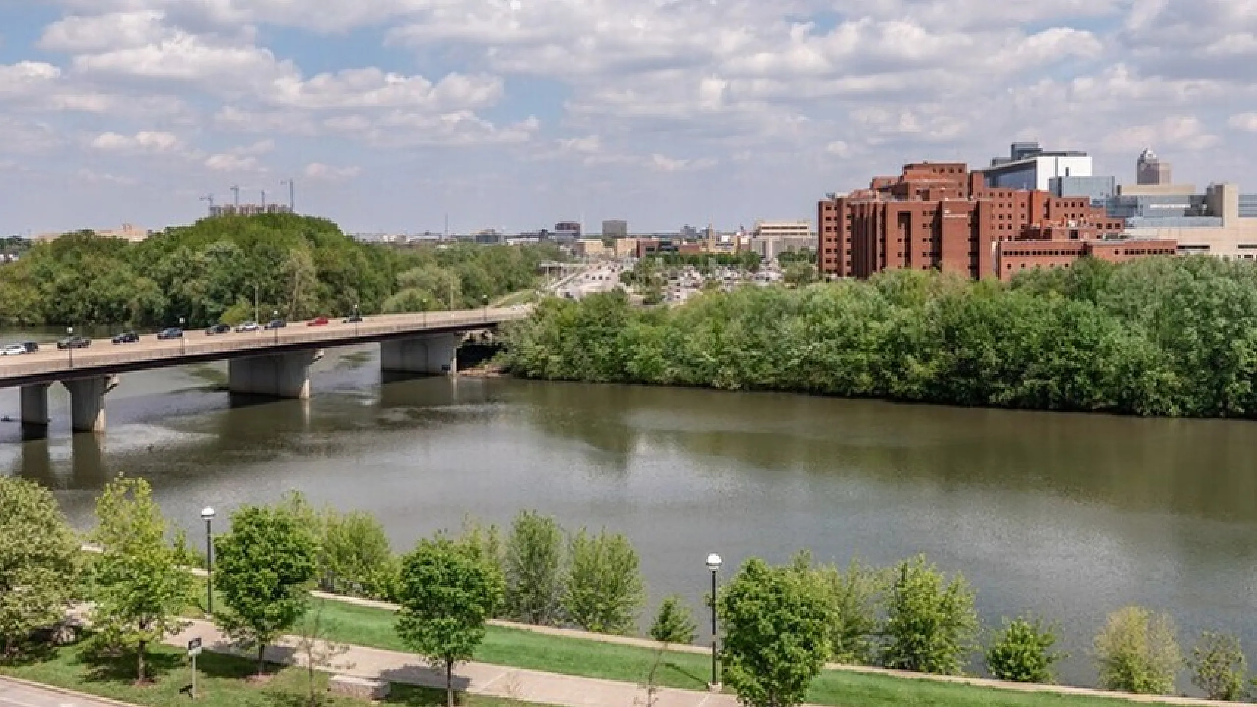 Scenic city view with a lush riverbank, iconic bridge, and modern skyline under a bright blue sky.