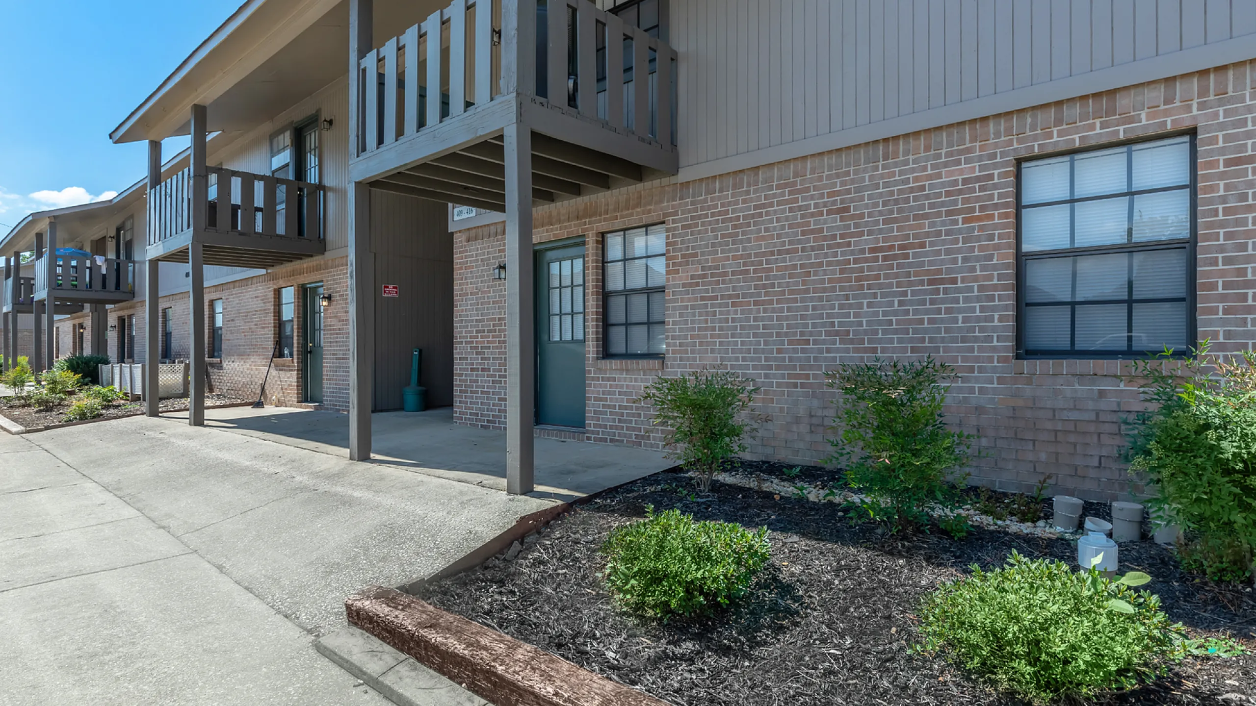 Charming apartment exterior featuring a brick facade, private balconies, and manicured landscaping under a clear blue sky.