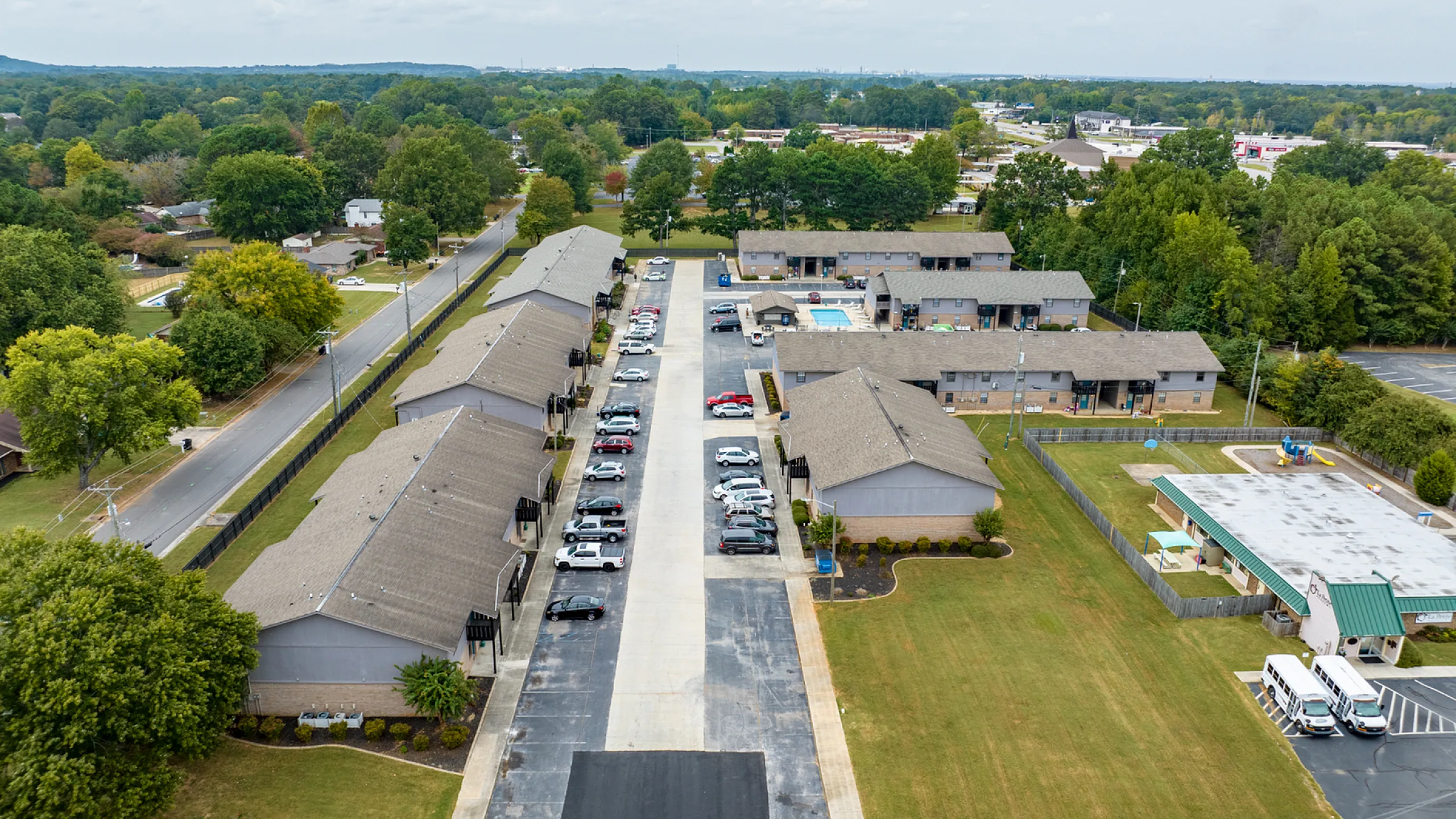 Aerial view of a charming apartment complex surrounded by lush greenery, featuring ample parking, a well-maintained play area, and community spaces in a peaceful suburban neighborhood.