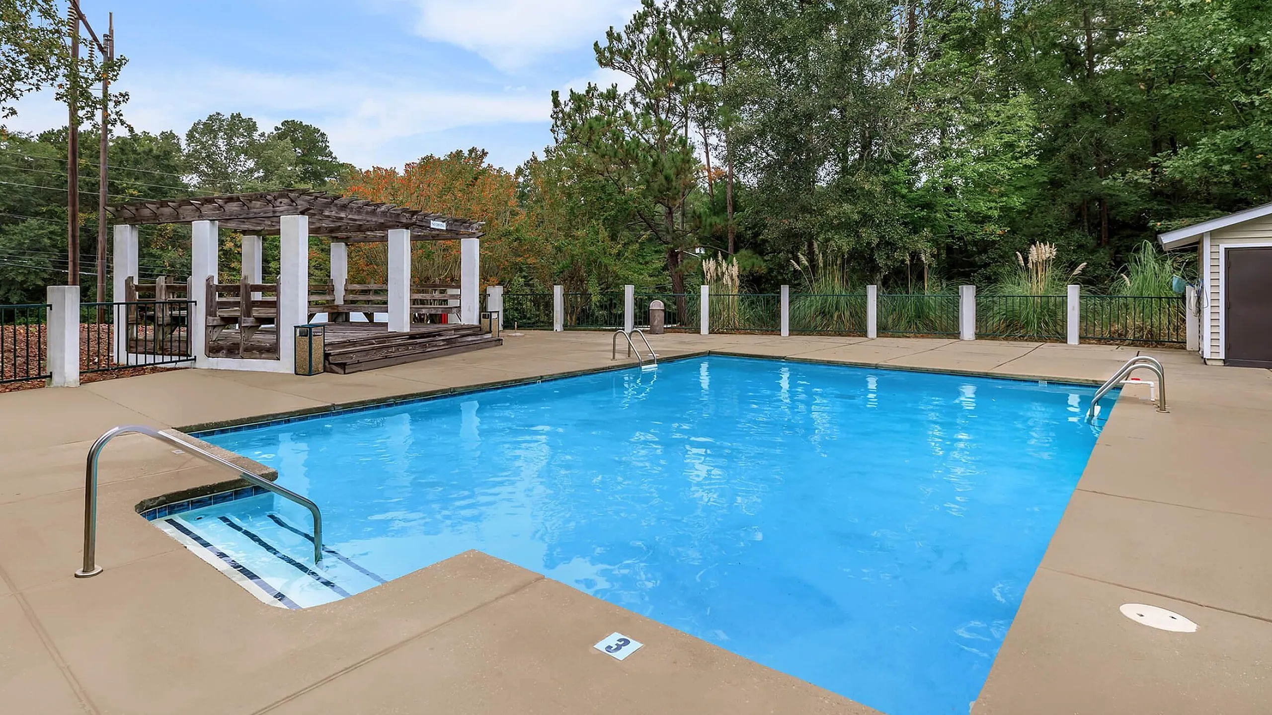 Inviting outdoor pool area with a spacious sundeck and charming pergola, surrounded by lush greenery for a tranquil retreat.