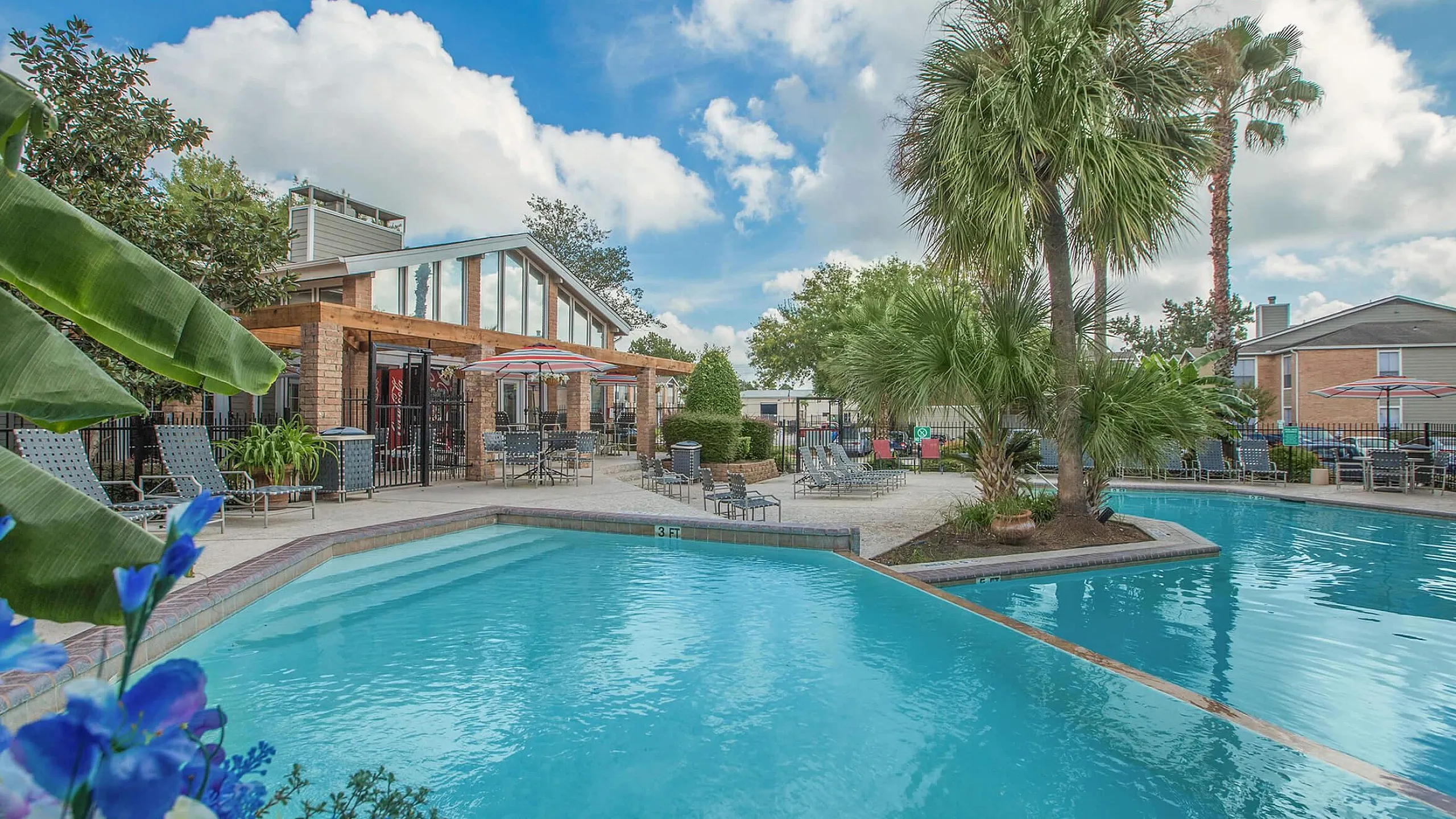 Inviting outdoor pool area with lush landscaping, comfortable seating, and a modern clubhouse under bright blue skies.