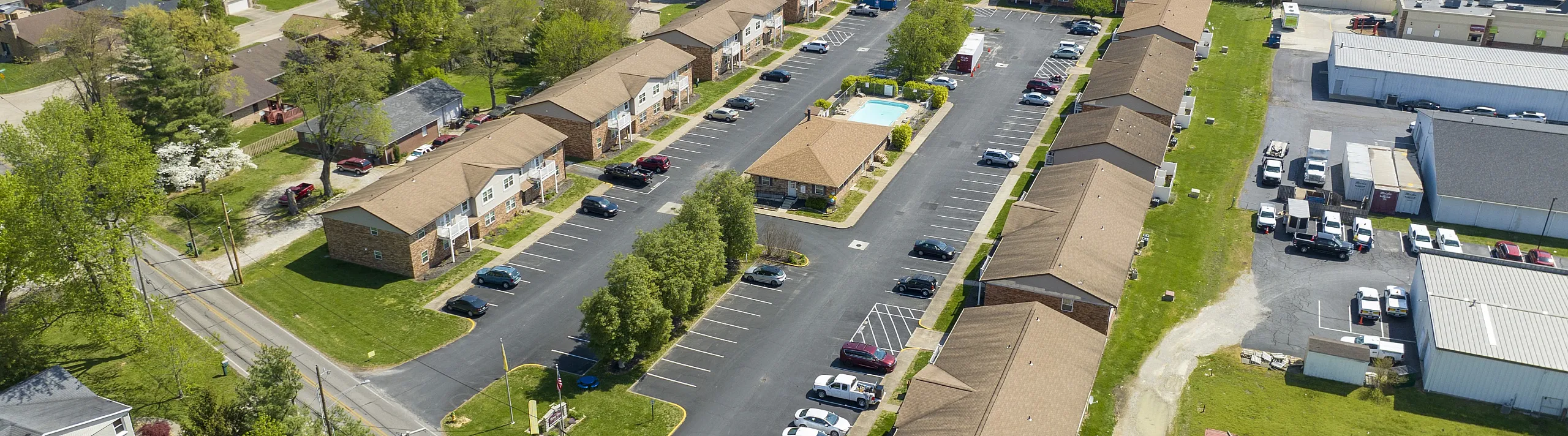 Aerial view of a charming apartment complex with ample parking, lush greenery, and a central swimming pool, surrounded by a vibrant community.