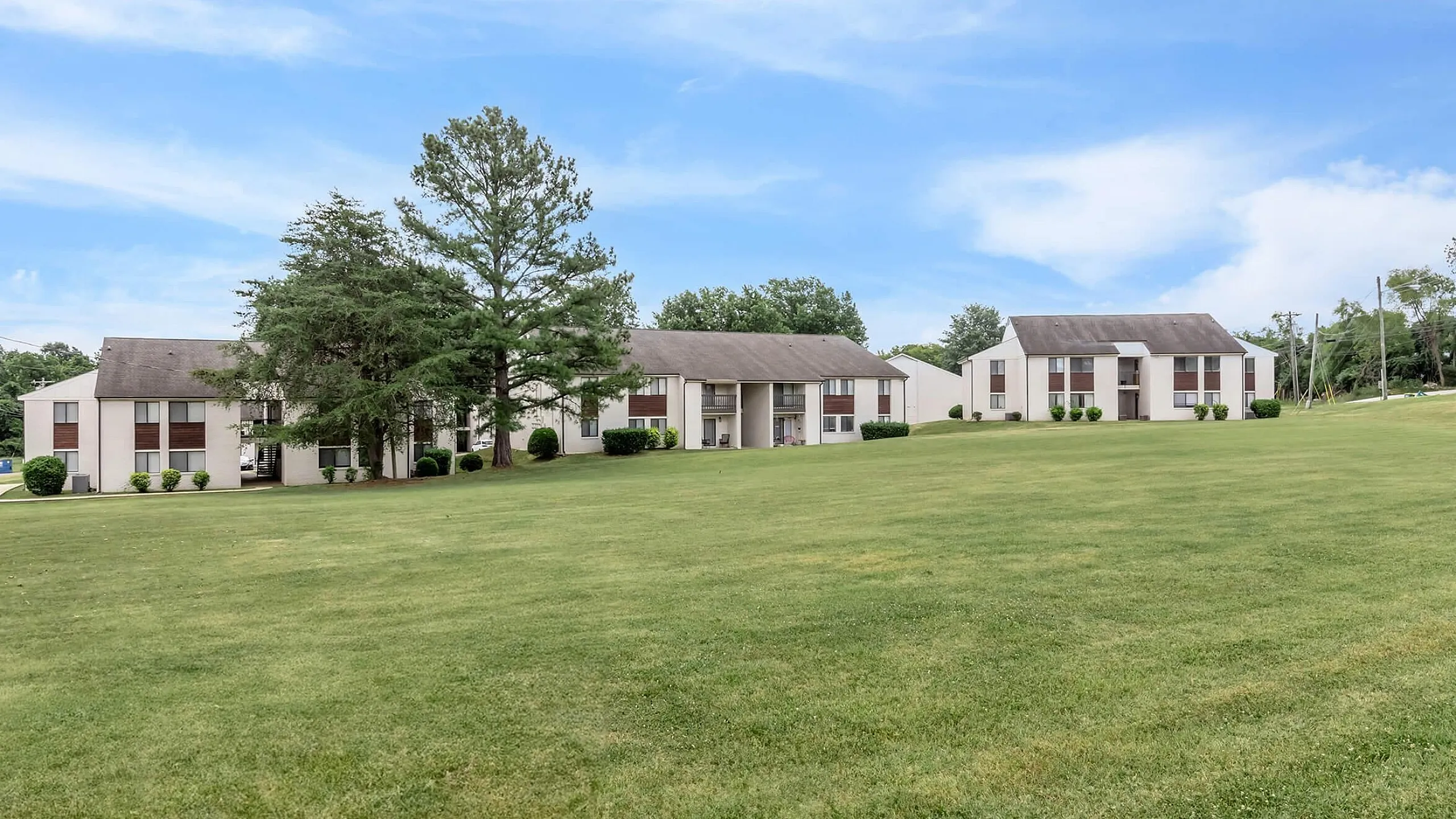 Inviting apartment community featuring charming white buildings surrounded by lush green lawns and mature trees under a bright blue sky.