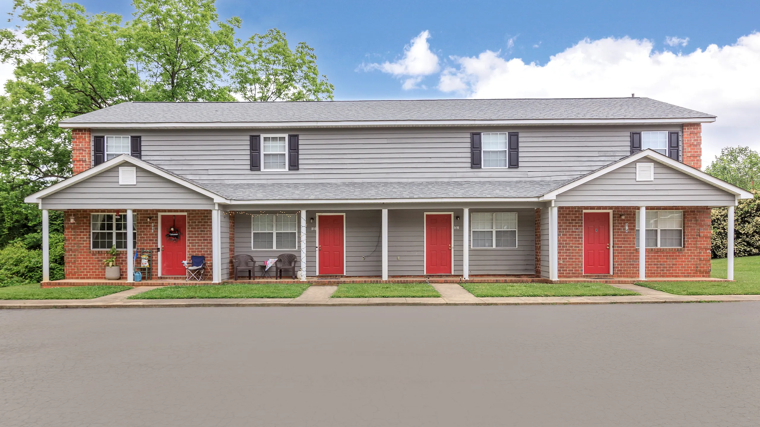 Charming duplex with red front doors and a cozy porch, set against a lush green backdrop.