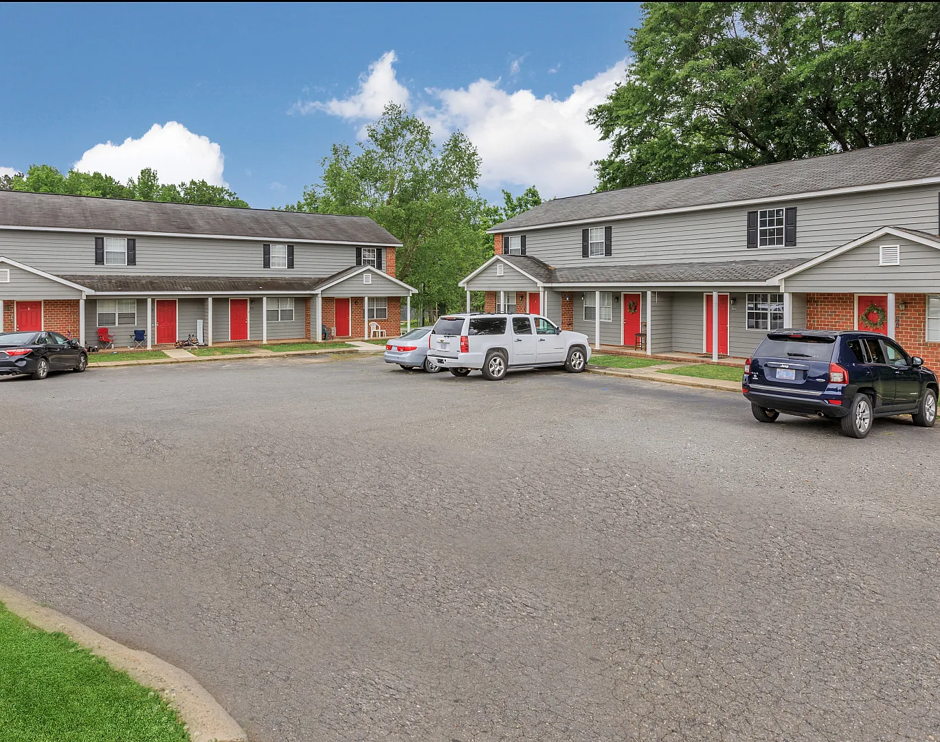 Charming two-story apartment complex with red doors, surrounded by lush greenery and ample parking space, under a clear blue sky.