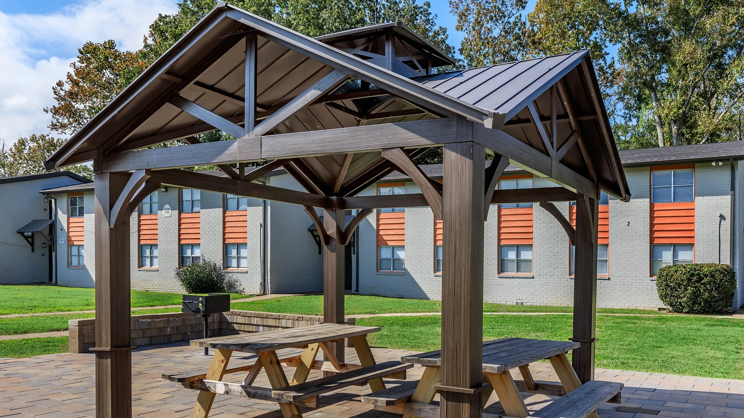 Charming outdoor patio with a cozy wooden gazebo and picnic tables, set in a vibrant green courtyard at a modern apartment community.