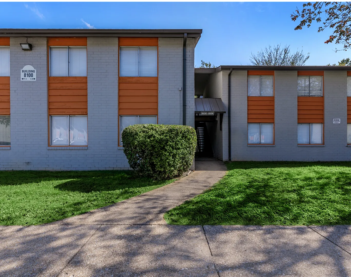 Charming apartment building with modern orange and gray brick facade, surrounded by lush green landscaping and a welcoming sidewalk entrance.
