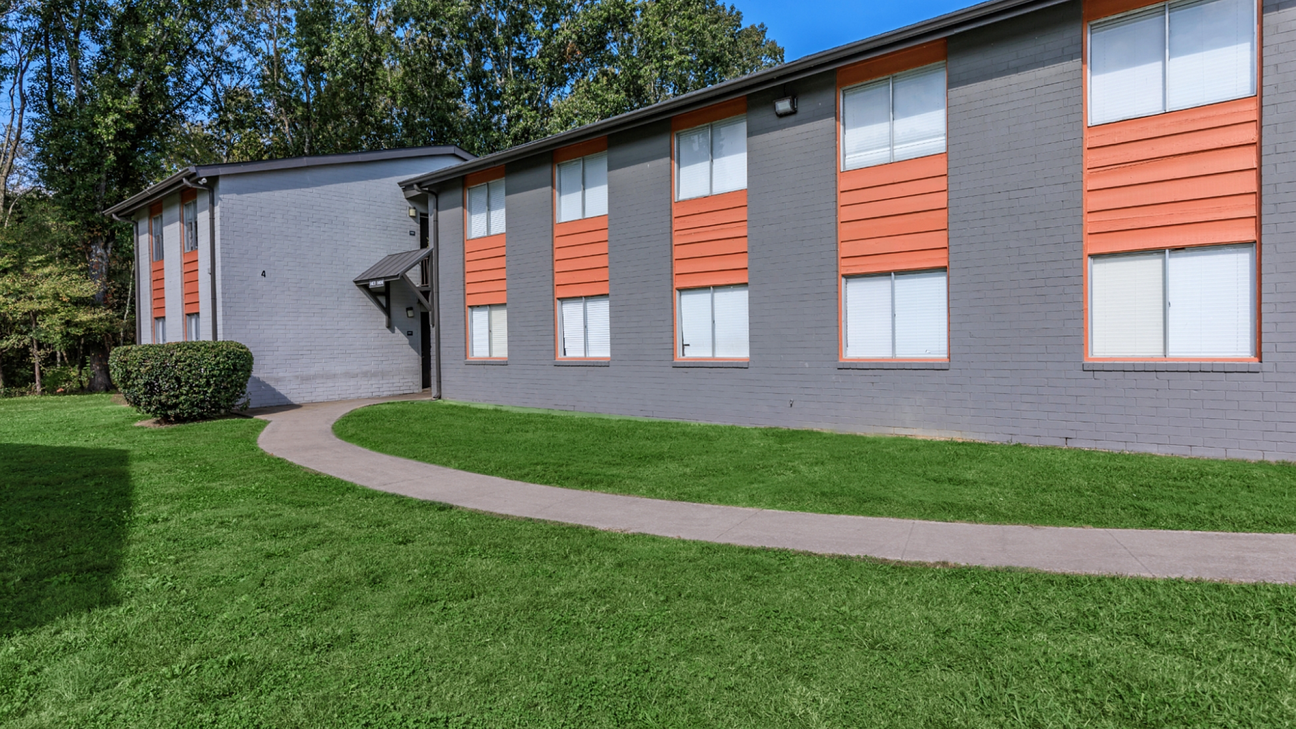 Charming apartment building exterior with modern gray and orange accents, surrounded by lush greenery and a welcoming pathway.