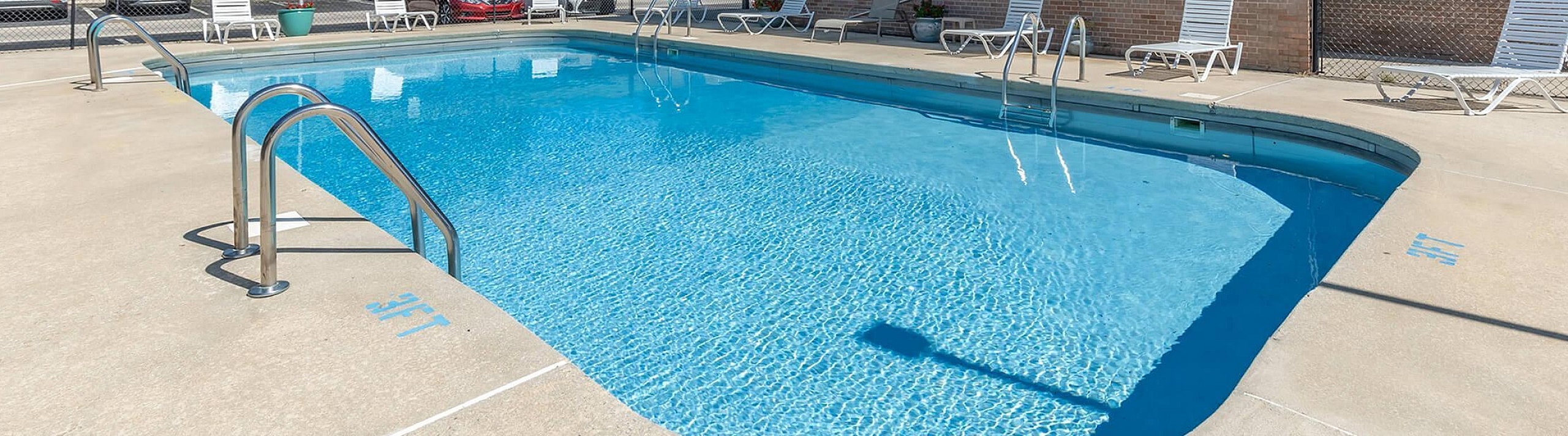 Sunny apartment complex with a sparkling blue swimming pool, surrounded by lounge chairs and lush greenery, perfect for relaxation.