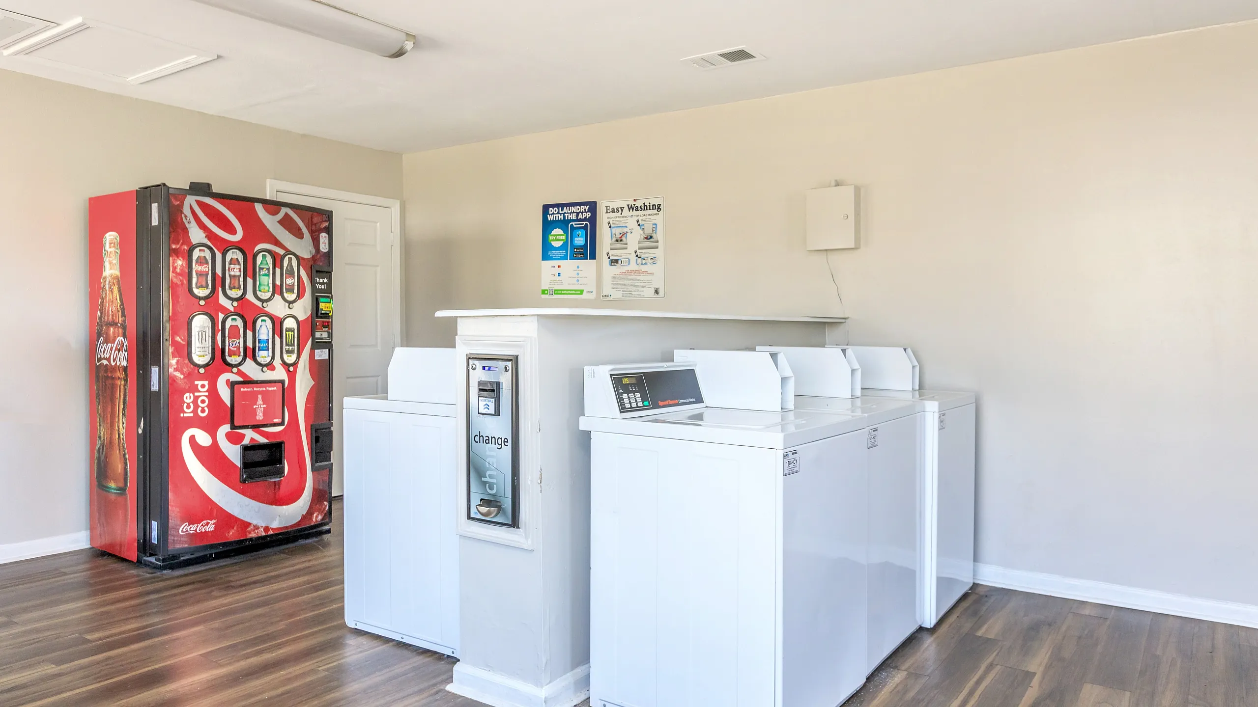 Bright, clean laundry room featuring modern white washing machines and a convenient beverage vending machine on sleek wooden flooring.