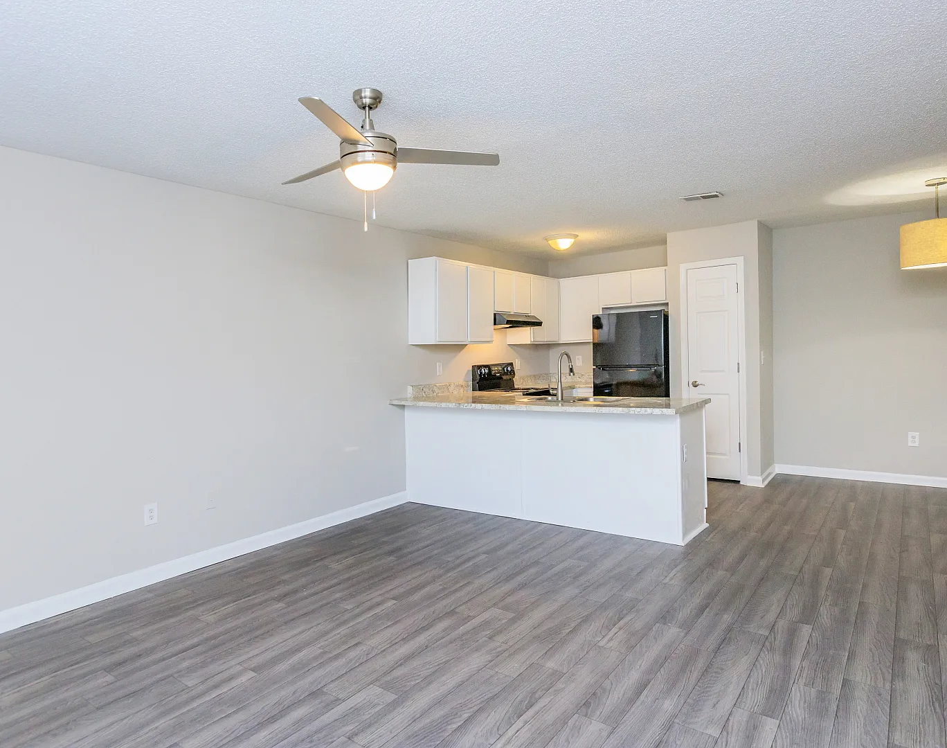 Modern open-plan living area featuring sleek wooden flooring, ceiling fan, and a stylish kitchen with white cabinetry and granite countertops.
