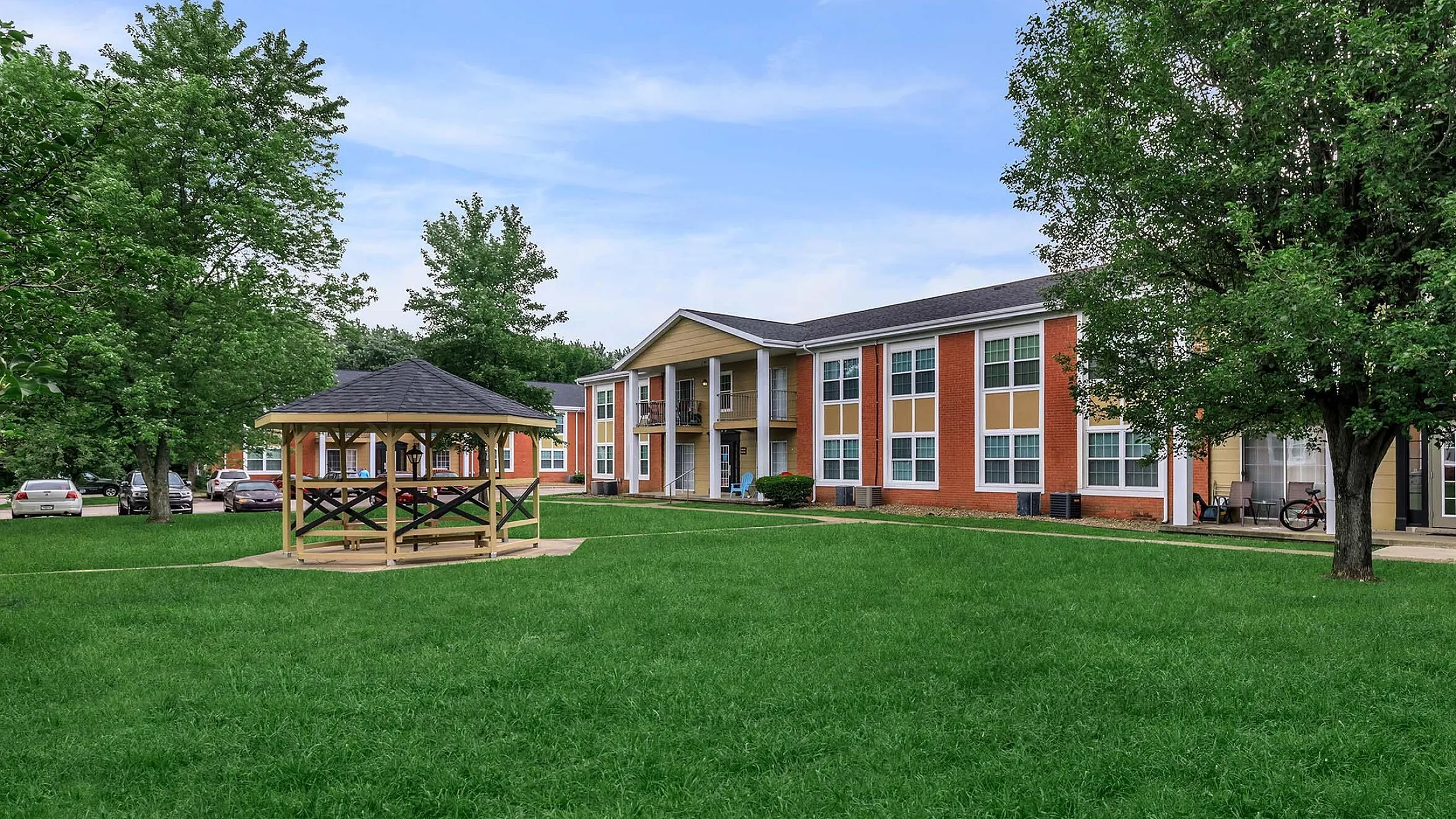 Charming two-story apartment complex with vibrant brick facade, lush green lawn, and a cozy wooden gazebo, perfect for community gatherings.