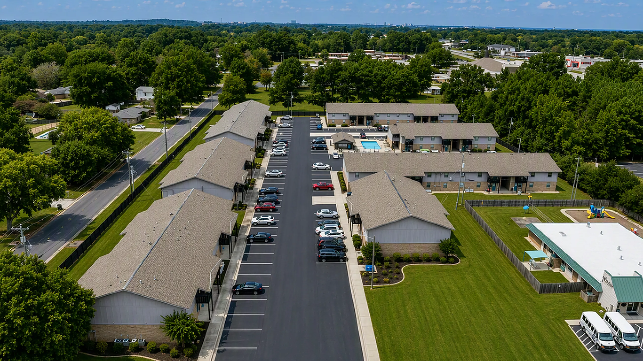 Aerial view of a charming apartment complex featuring neatly arranged buildings, ample parking, lush green lawns, and a refreshing outdoor pool, set against a serene suburban backdrop.