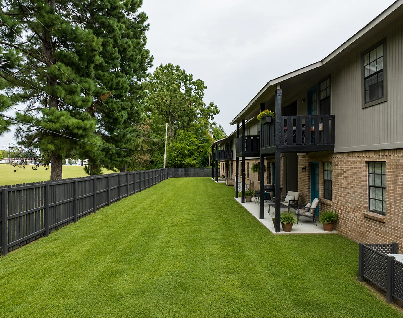 Charming apartment exterior with brick facade, overlooking a lush green lawn and cozy outdoor seating areas, surrounded by tall pine trees.