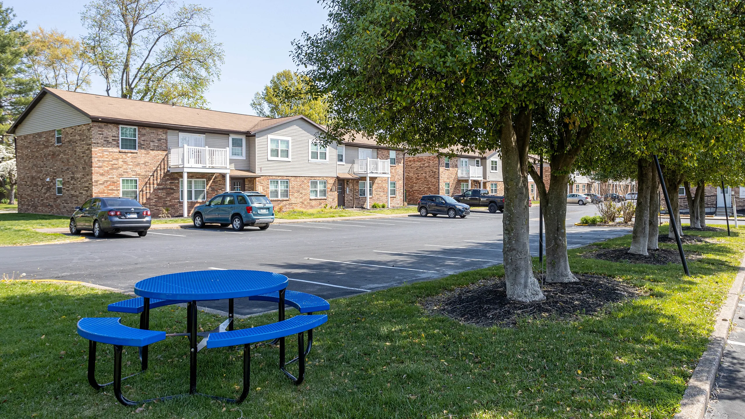Charming apartment complex surrounded by lush green trees, featuring ample parking and a vibrant blue picnic area for outdoor relaxation.