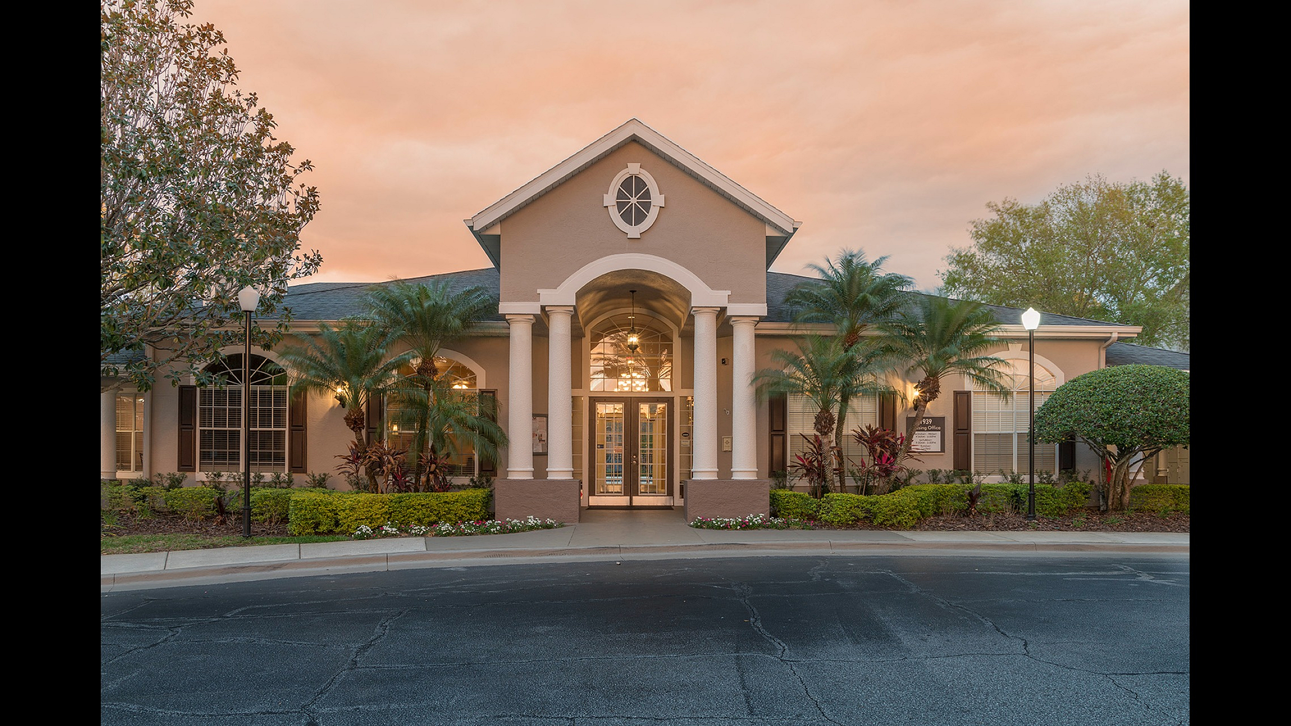 Elegant apartment entrance with grand columns, lush landscaping, and inviting evening lighting under a picturesque sunset.