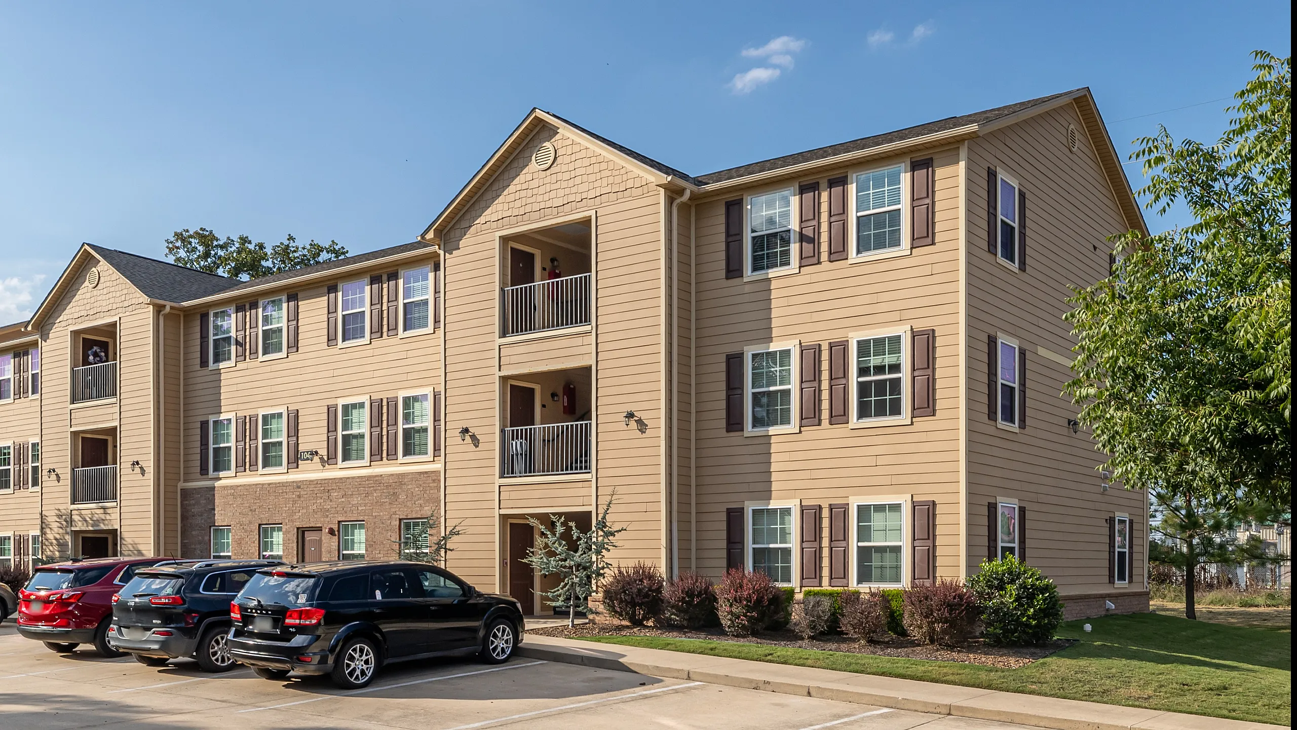 Charming multi-story apartment building with ample parking and attractive landscaping under a bright blue sky.