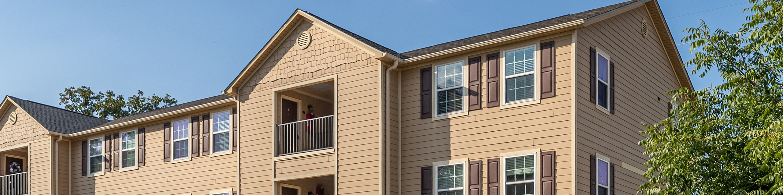 Charming three-story beige apartment building with stylish shutters and ample parking spaces, set against a clear blue sky and surrounded by lush greenery.