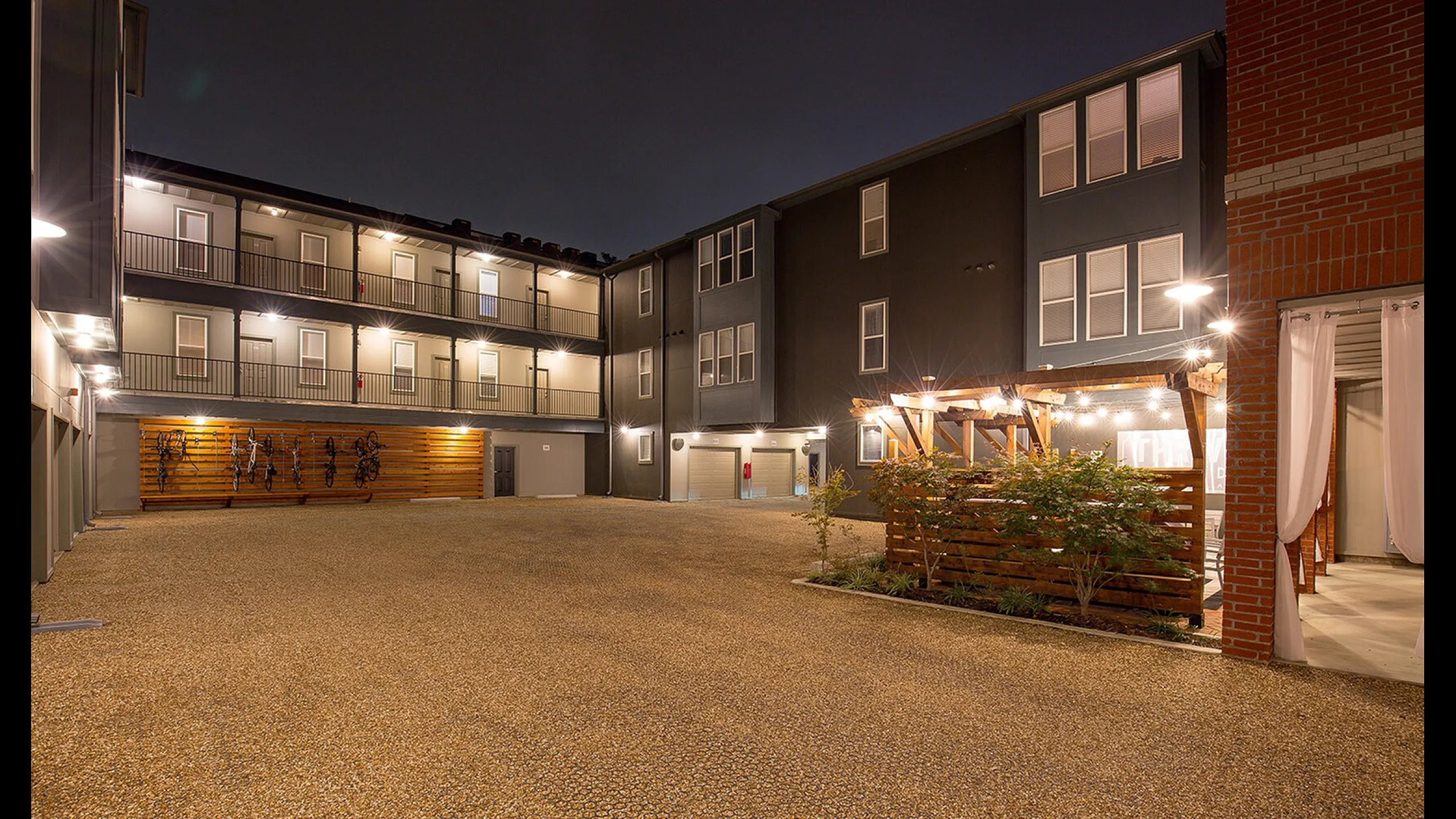 Charming modern apartment courtyard at night featuring well-lit walkways, bicycle racks, and a wooden pergola for relaxation.