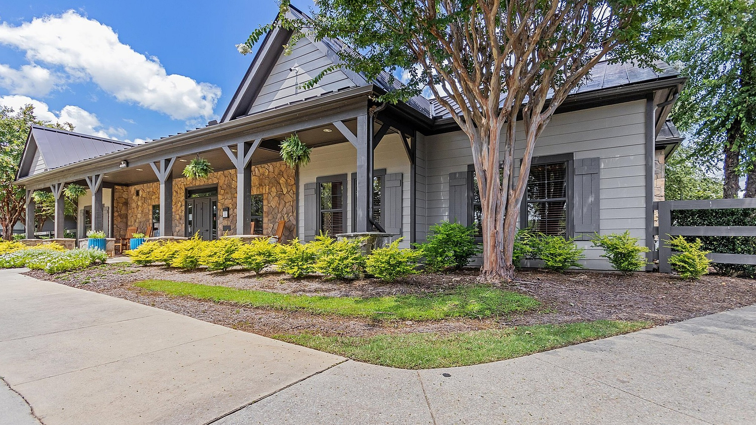 Charming residential building with a stone facade and inviting front porch surrounded by lush greenery under a vibrant blue sky.