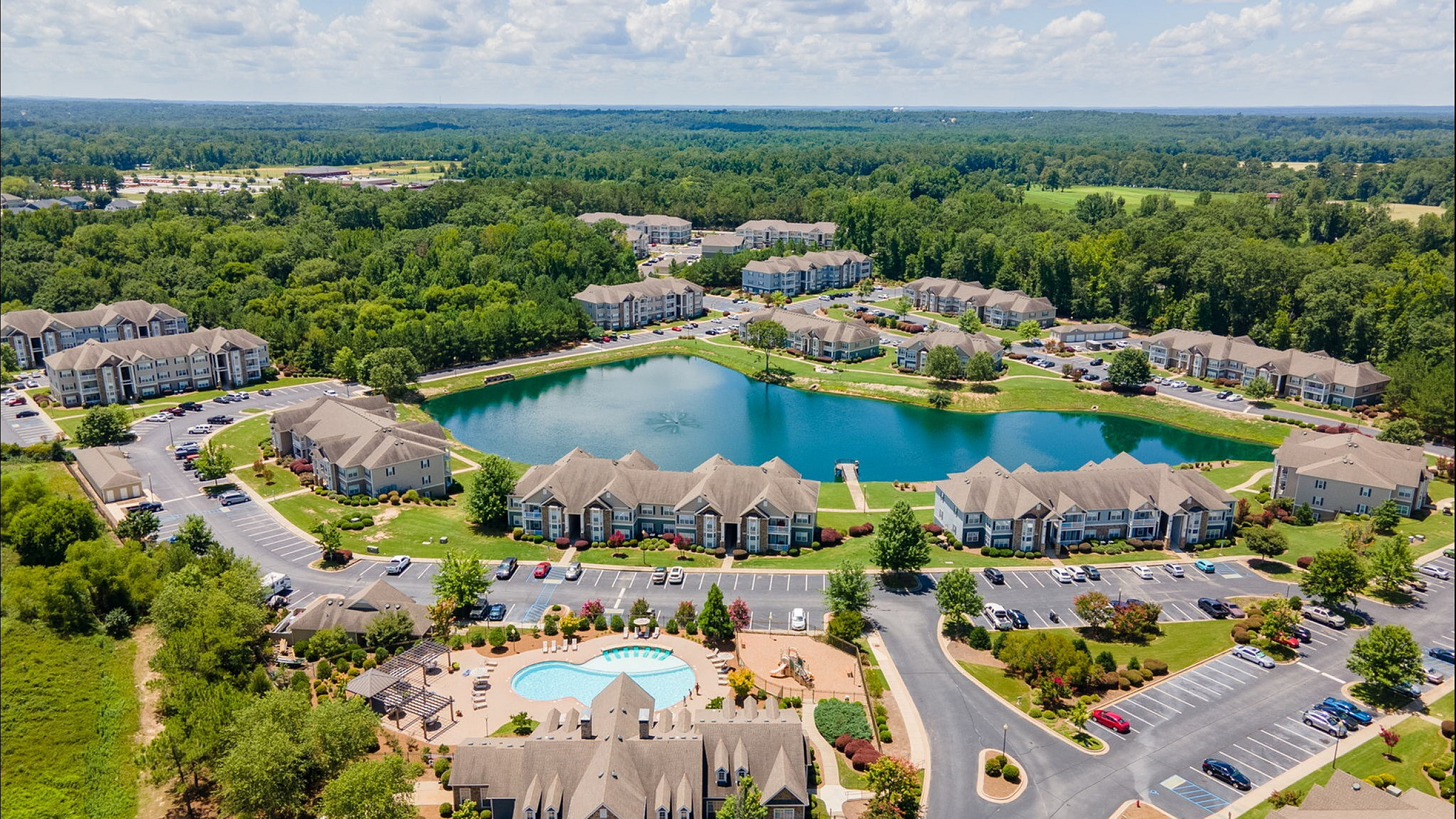 Aerial view of a charming apartment community featuring a picturesque lake, spacious green spaces, and a refreshing pool area surrounded by modern buildings.