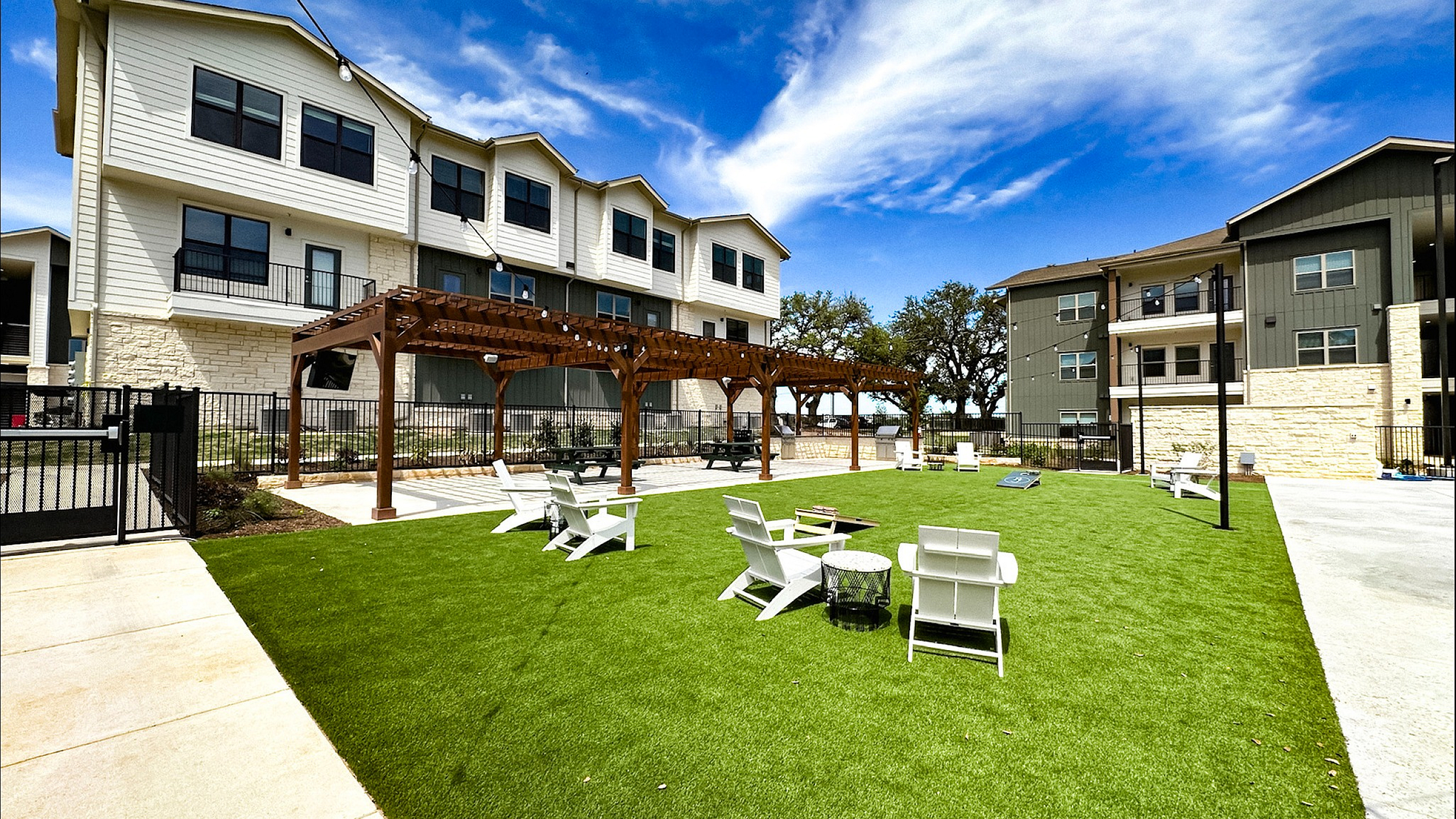 Charming apartment complex with inviting outdoor space, featuring a lush green lawn, stylish pergola, and cozy seating under a clear blue sky.