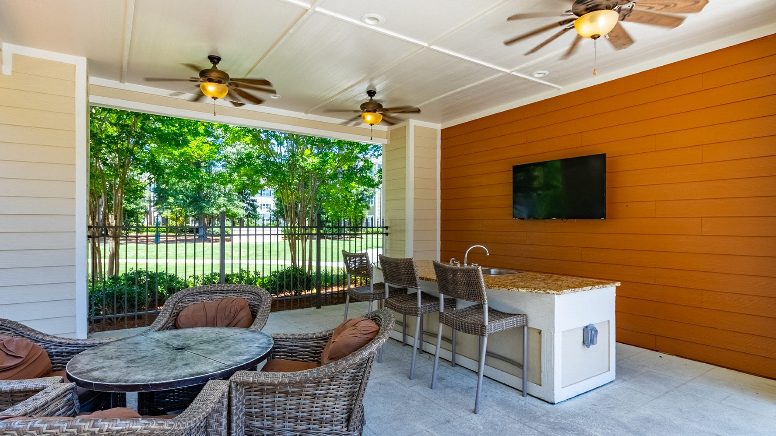 Charming outdoor seating area with wicker chairs and a TV, shaded by ceiling fans, perfect for relaxing while enjoying garden views.