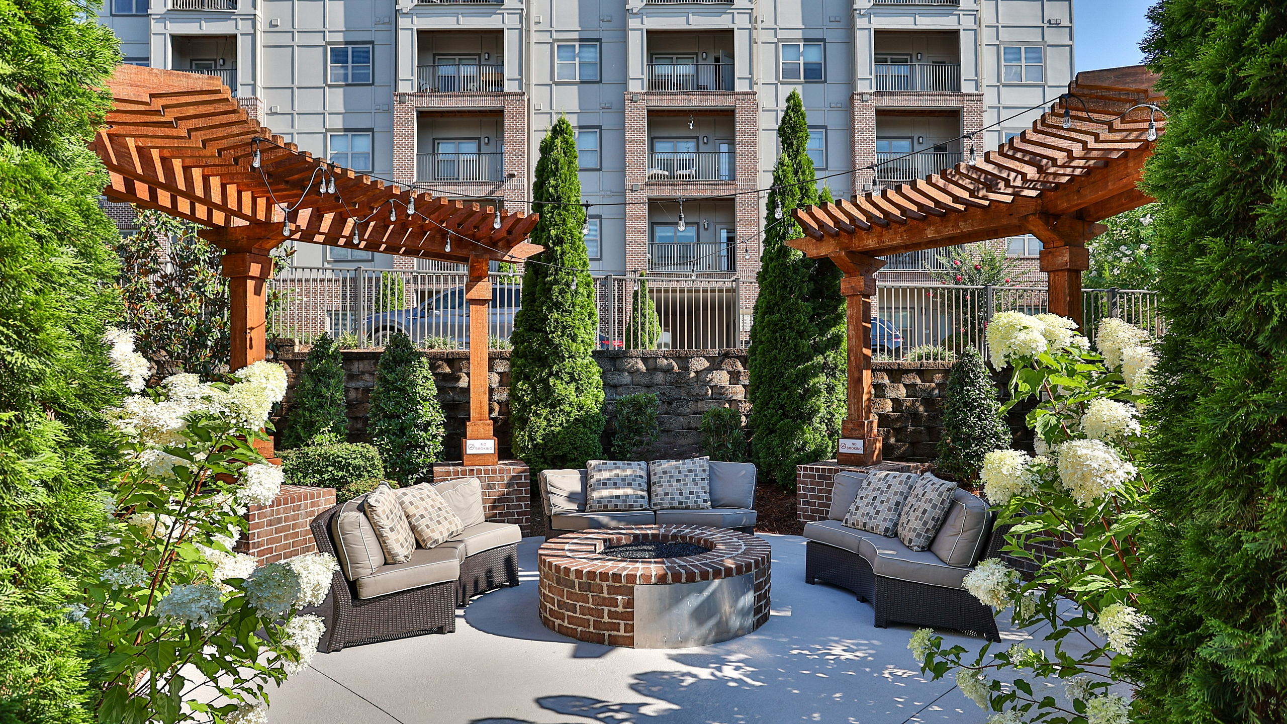 Charming outdoor seating area with cozy sofas, a brick fire pit, and elegant wooden pergolas, set against a stylish apartment building backdrop.