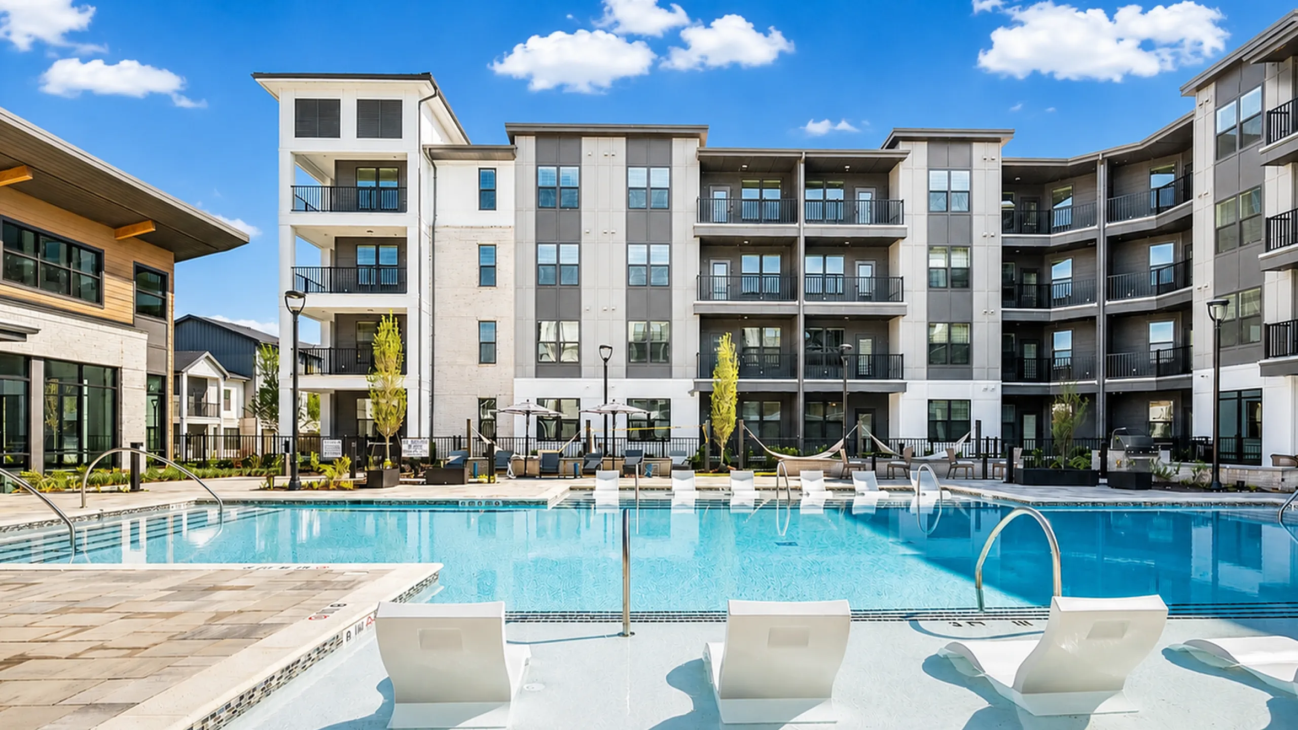 Luxurious apartment complex with a pristine outdoor pool, stylish lounge chairs, and modern architecture under a clear blue sky.