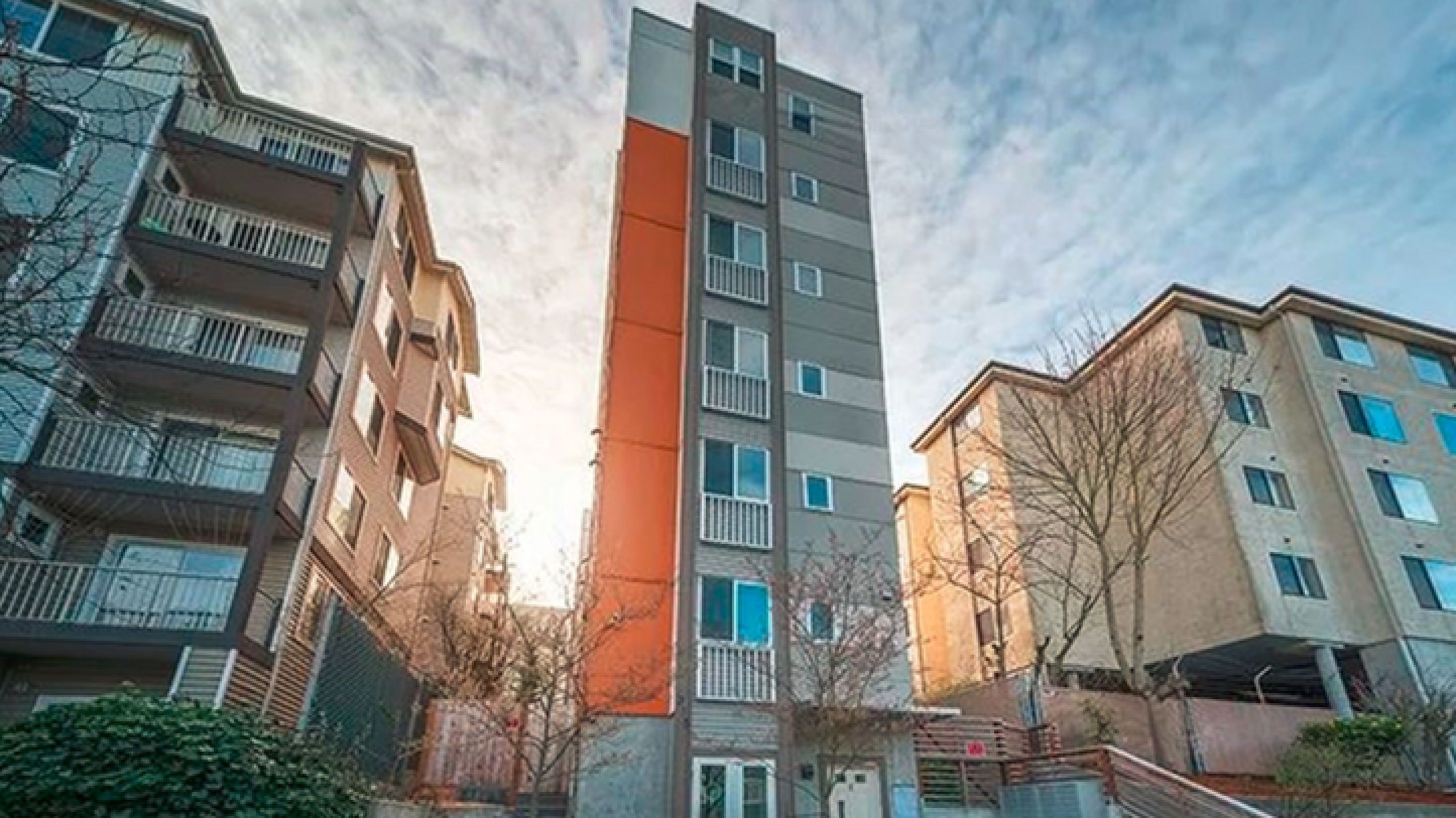 Modern apartment building with striking orange accents, surrounded by lush greenery and a welcoming path in a vibrant urban neighborhood.