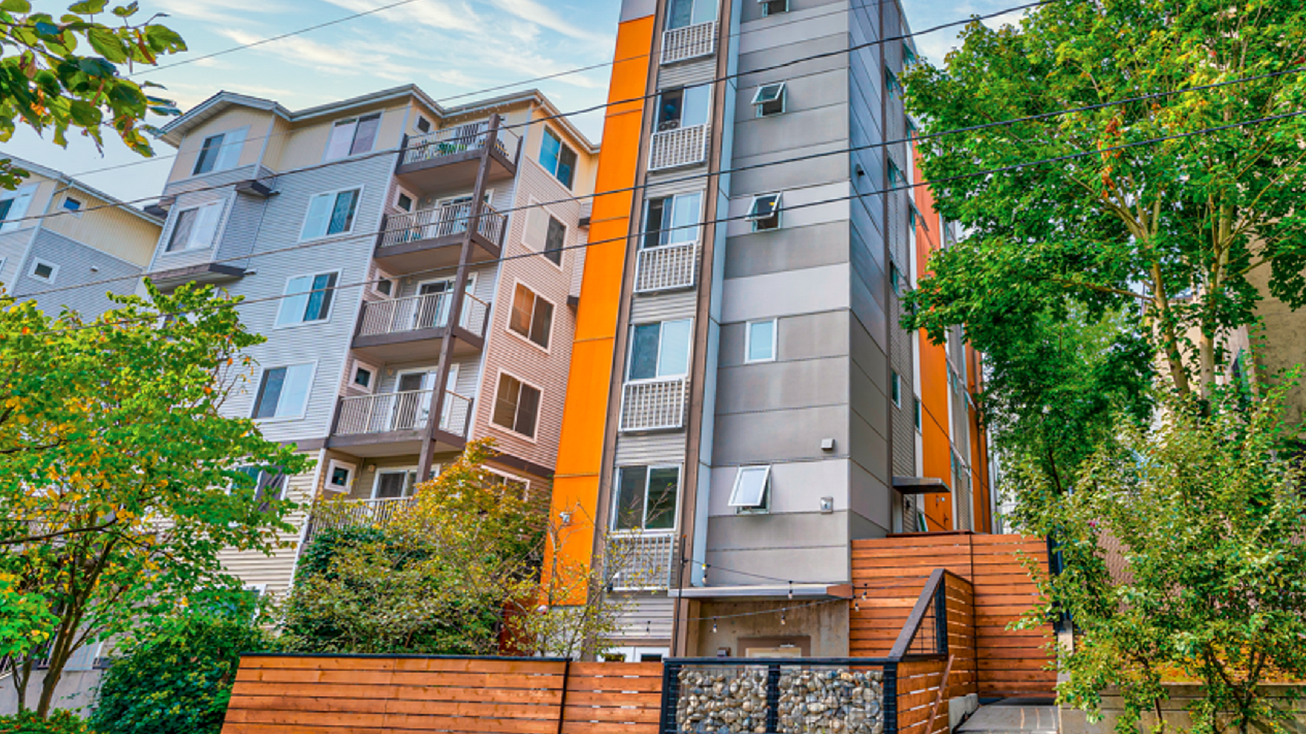 Modern apartment building with vibrant orange accents, surrounded by lush greenery and a clear blue sky.