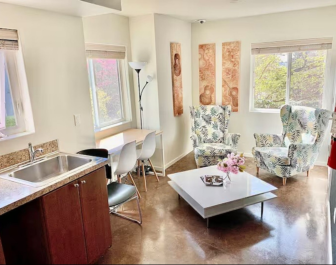 Bright and inviting living space with chic floral armchairs, large windows, elegant coffee table, and a cozy dining nook adjacent to the kitchen.