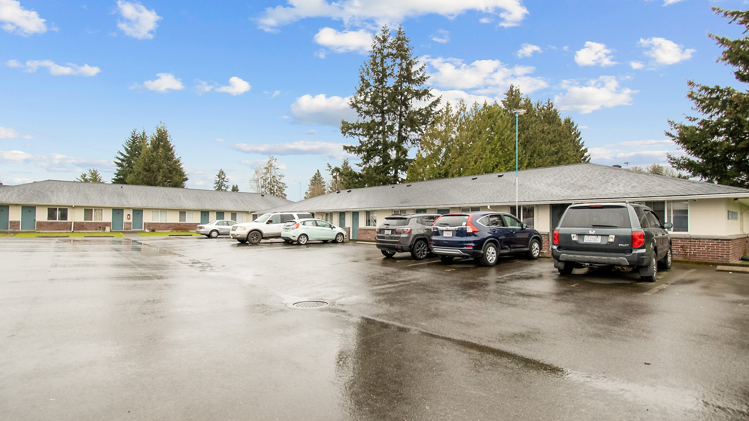 Charming single-story apartment complex with ample parking and surrounded by lush green trees under a vibrant blue sky.