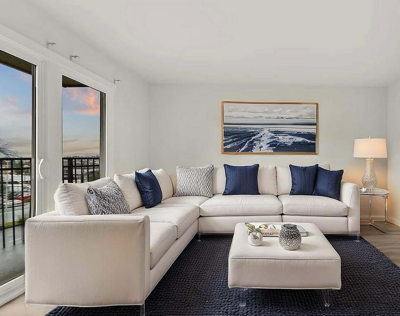 Contemporary living room featuring a large white sectional sofa with navy and patterned cushions, cozy ambiance with a chic lamp, modern art piece, and a wide flat-screen TV; expansive windows offer stunning views.