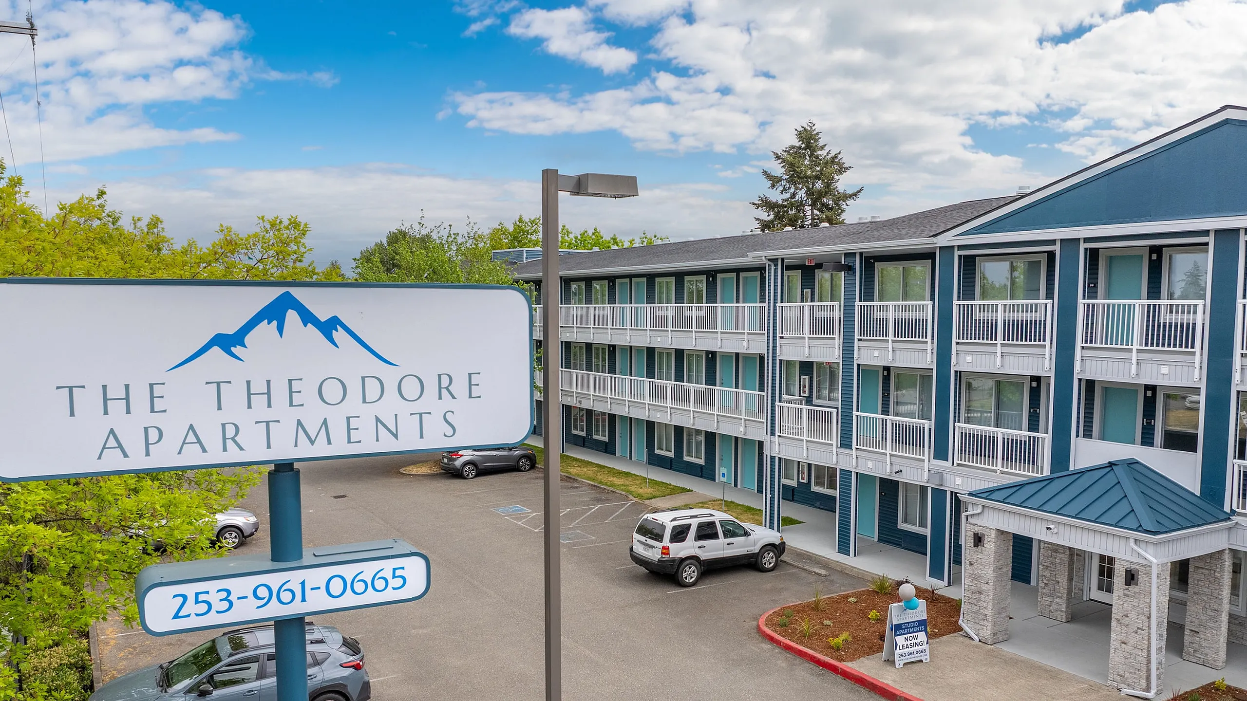Stunning view of The Theodore Apartments featuring modern architecture and ample parking space under a clear blue sky.