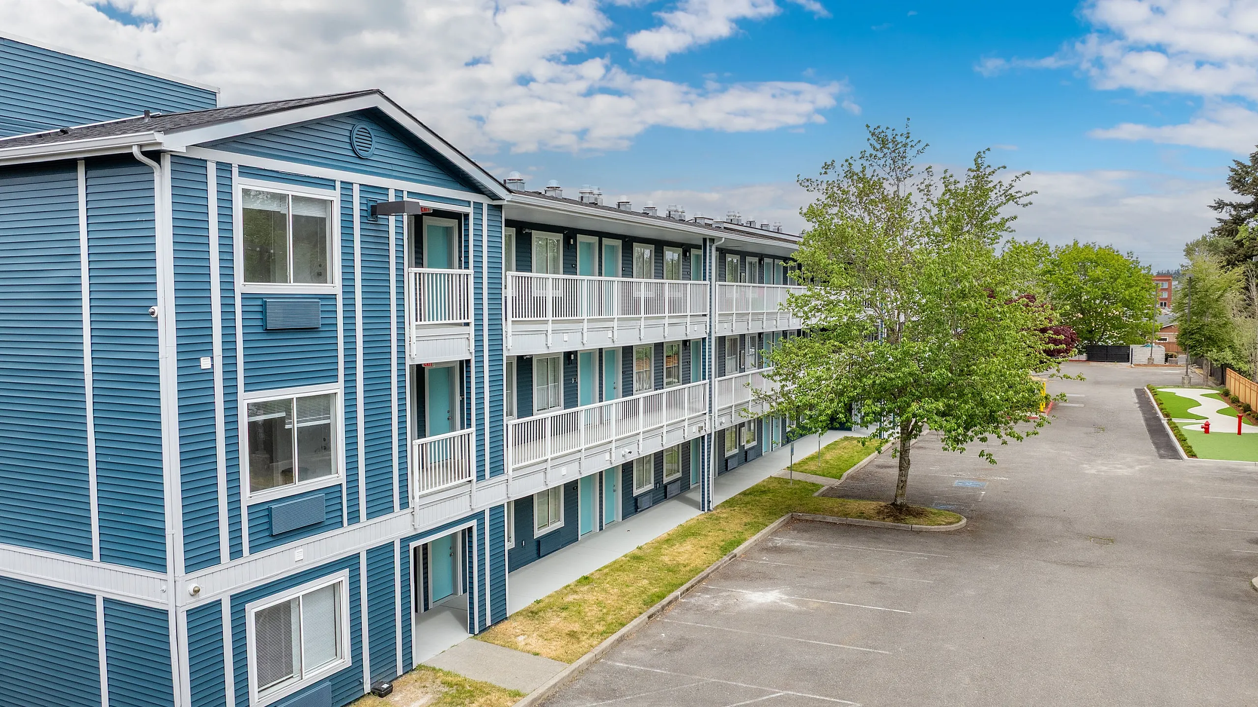 Charming blue apartment complex with elegant white accents, ample parking, and lush green surroundings under a vibrant blue sky.